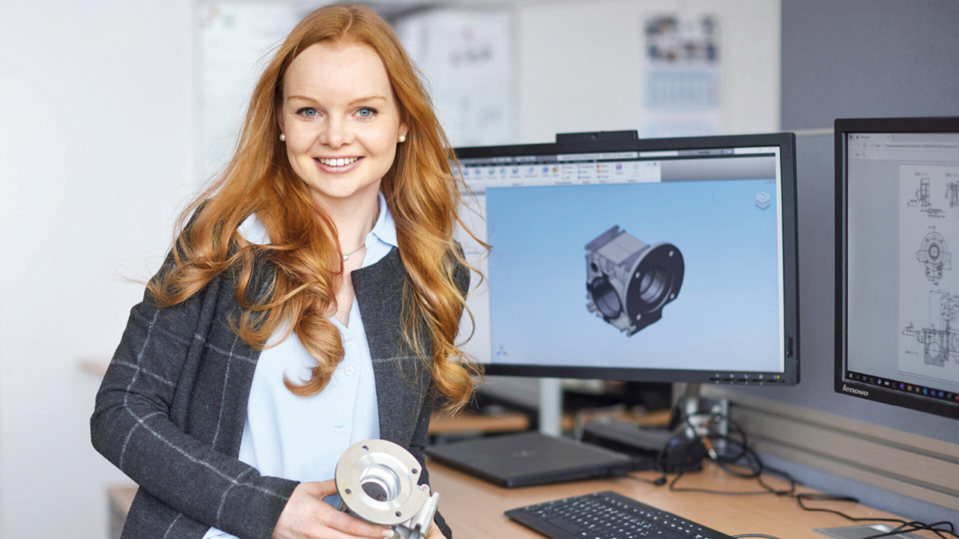 Eine lächelnde Frau hält ein mechanisches Teil und steht neben einem Schreibtisch mit zwei Monitoren, auf denen mechanische 3D-Konstruktionen in einem Büro oder einem technischen Arbeitsbereich angezeigt werden.