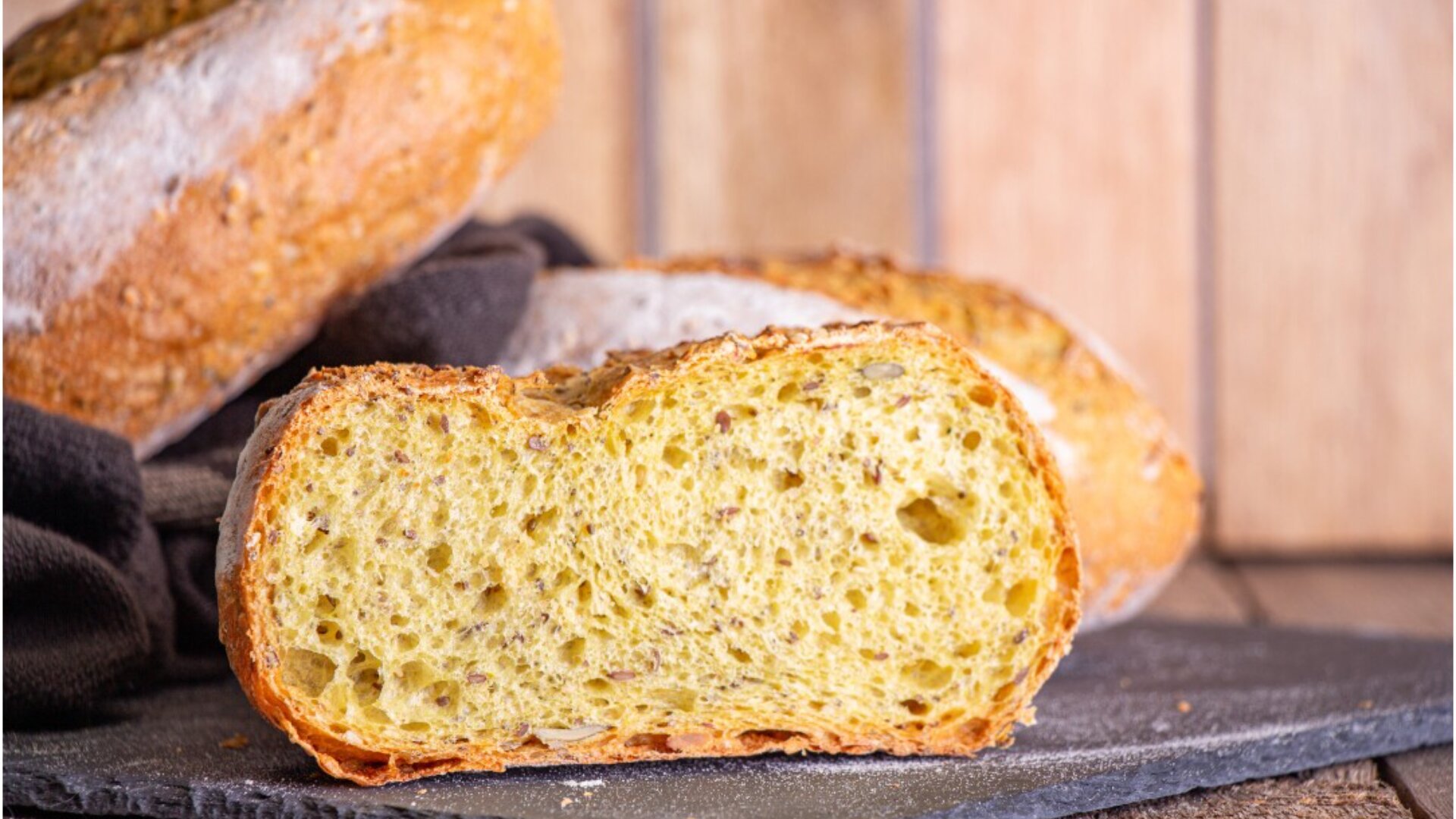 Ein aufgeschnittener Laib rustikalen Brotes mit einem goldenen, luftigen Inneren ruht auf einem dunklen Tuch und einer Steinplatte, mit einem weiteren ungeschnittenen Laib und einem hölzernen Hintergrund im Weichzeichner.