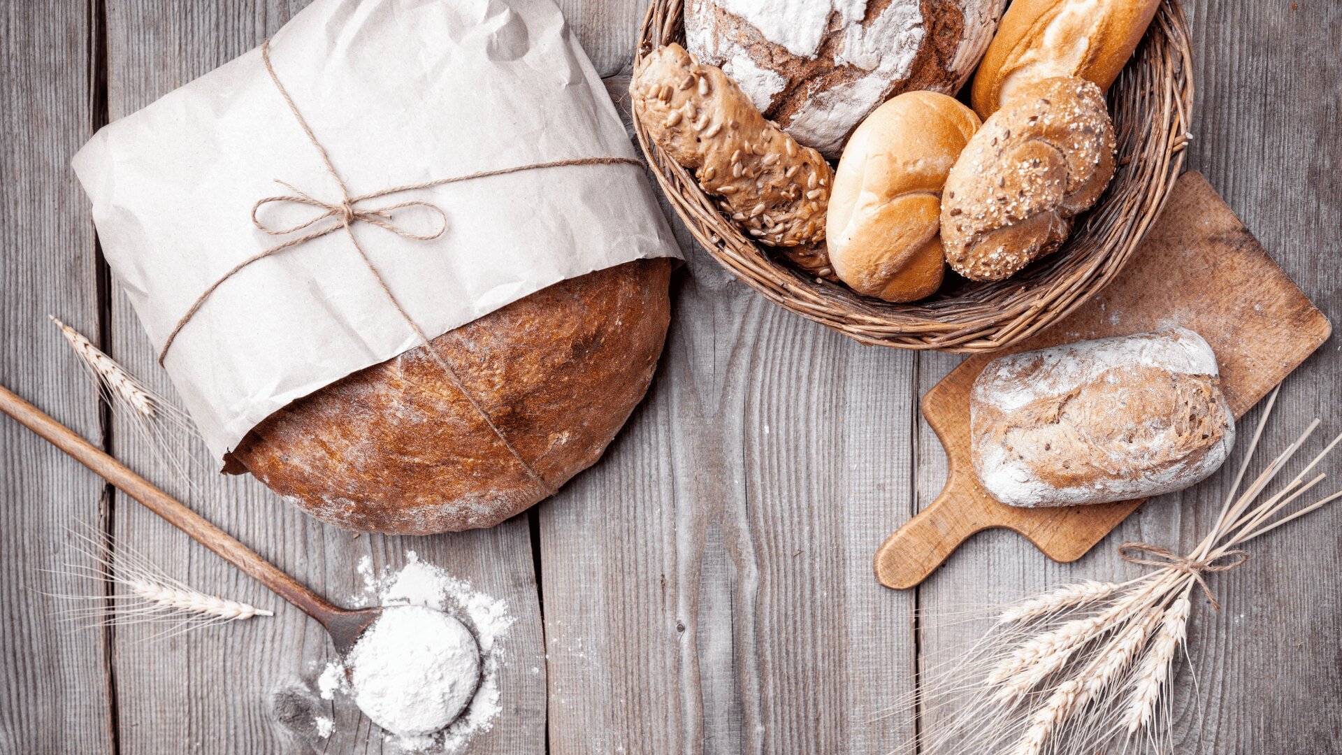 Ein in Papier eingewickelter Laib Brot, ein Korb mit verschiedenen Brötchen, ein Holzbrett mit einem kleinen Laib, ein Löffel Mehl und Weizenhalme auf einem rustikalen Holztisch.