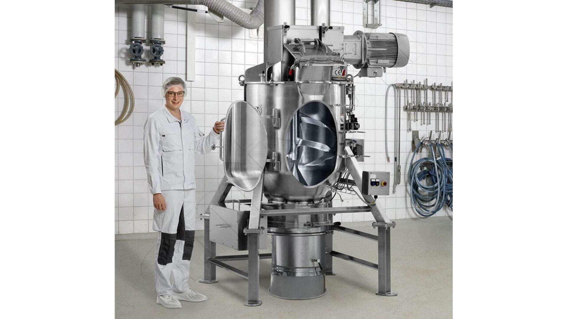 A person in a white lab coat and hair net stands beside a large industrial stainless steel mixing machine in a tiled room, with pipes and hoses visible in the background.