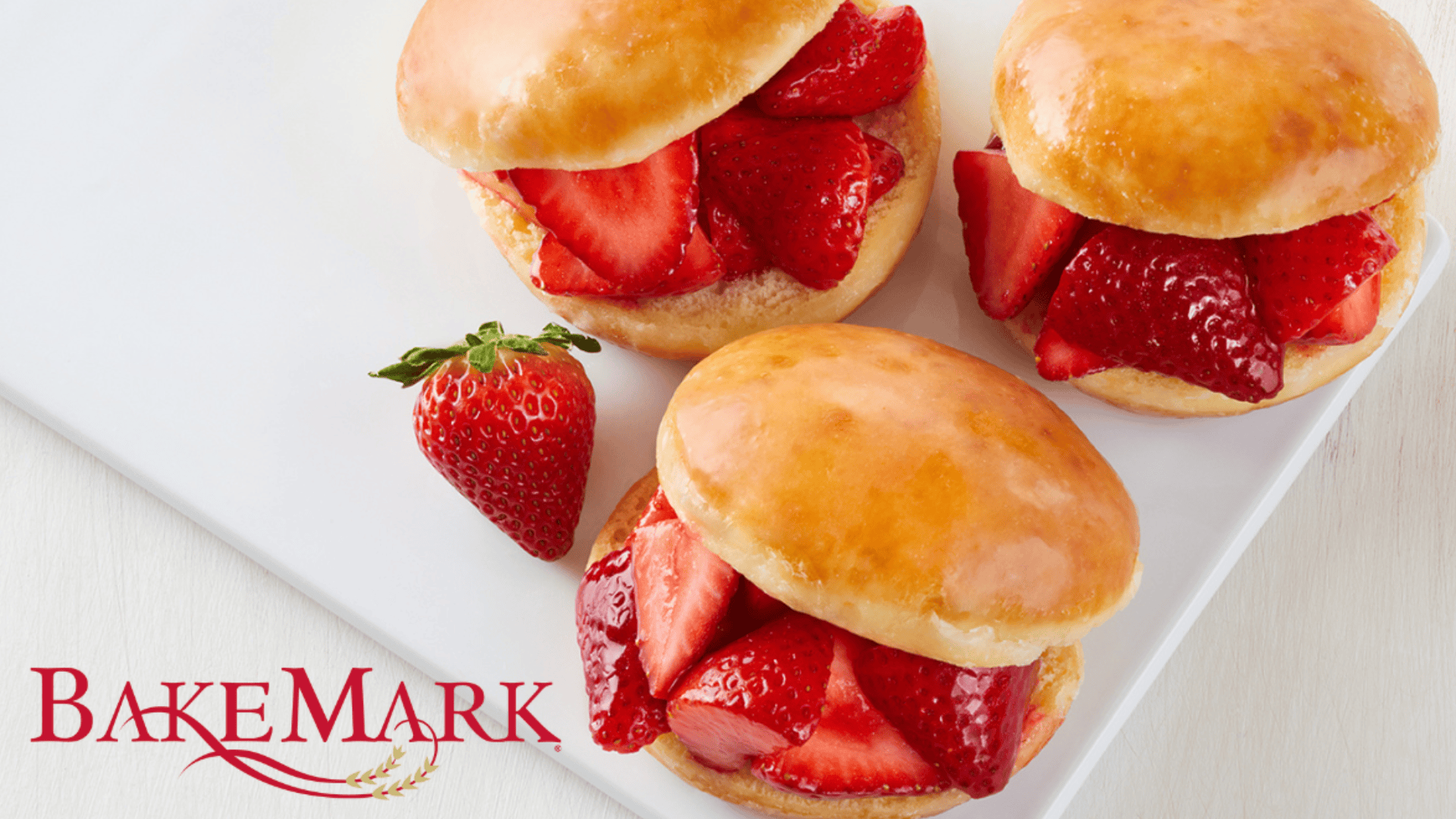Three glazed sweet buns filled with fresh sliced strawberries are arranged on a white plate next to a whole strawberry. The BakeMark logo appears in the lower left corner.