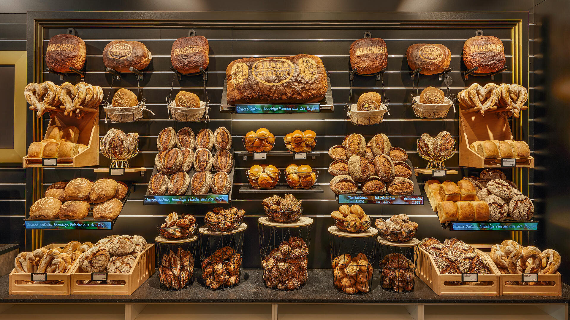 Eine Bäckereiauslage mit einer Vielzahl von Brotsorten und Brezeln, darunter große Brote, Brötchen und geflochtenes Brot, alles ordentlich auf Regalen und Tabletts vor einem dunklen Hintergrund angeordnet.