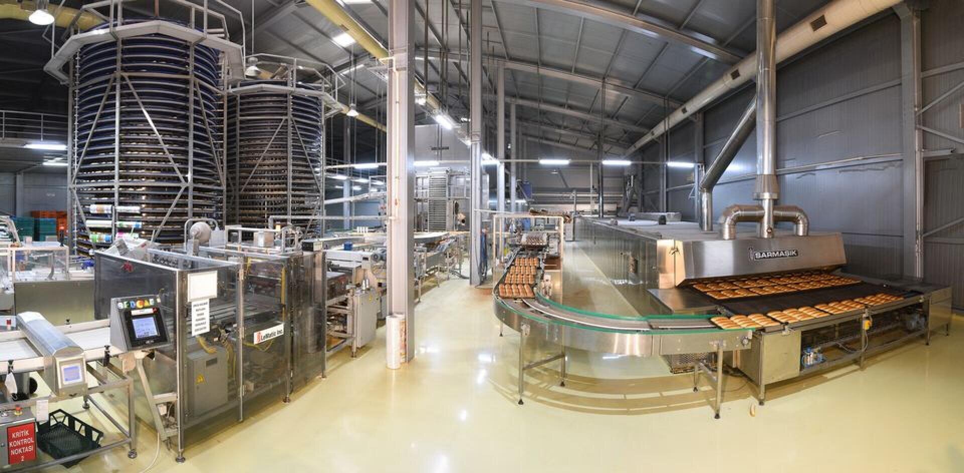 A modern industrial bakery with automated machines and conveyor belts producing and transporting baked goods, surrounded by large metal structures and high ceilings under bright lighting.