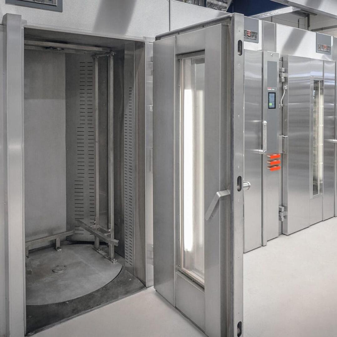 Ein großer Industrieofen aus rostfreiem Stahl mit einer offenen Tür, die den Blick auf eine kreisförmige Drehplattform im Inneren freigibt; daneben stehen geschlossene Öfen in einer sauberen, modernen Anlage.