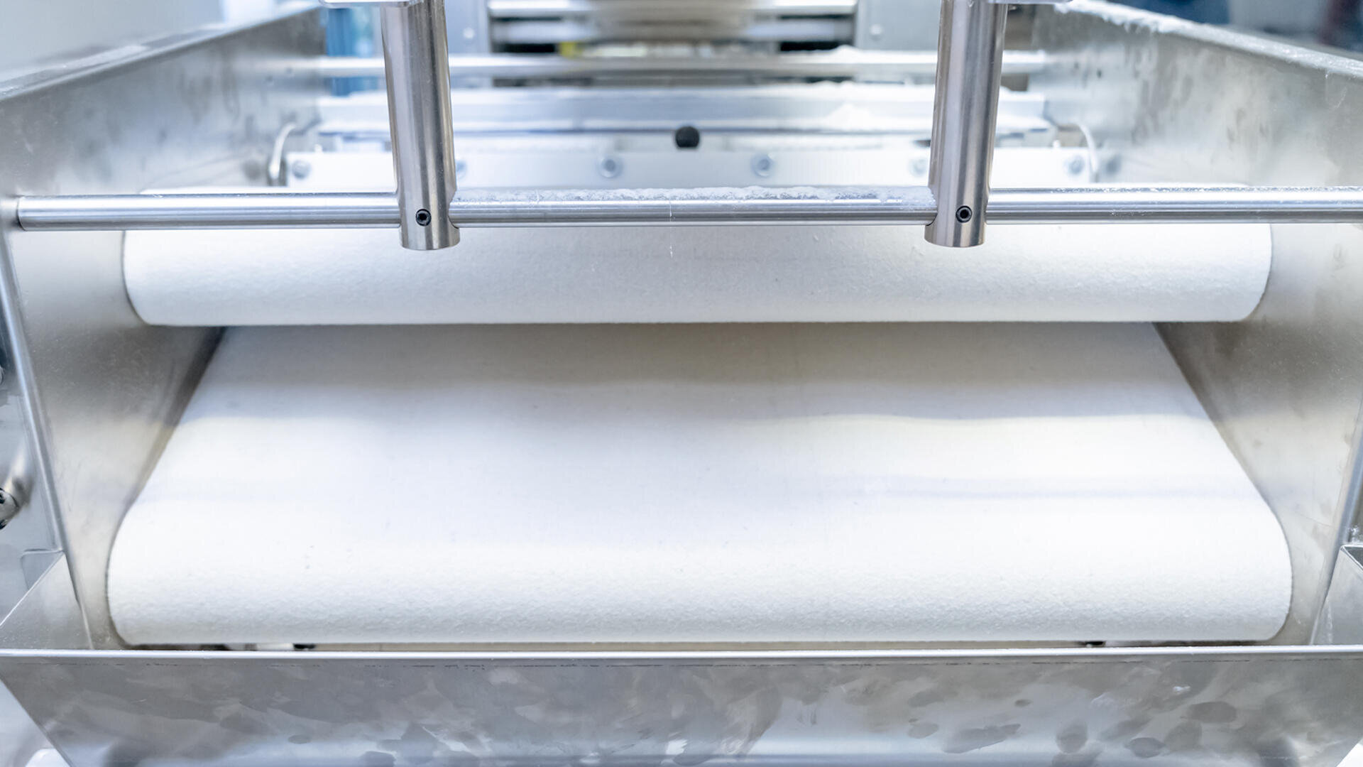 Close-up of an industrial dough sheeter machine flattening a sheet of white dough between two large metal rollers in a food processing facility.