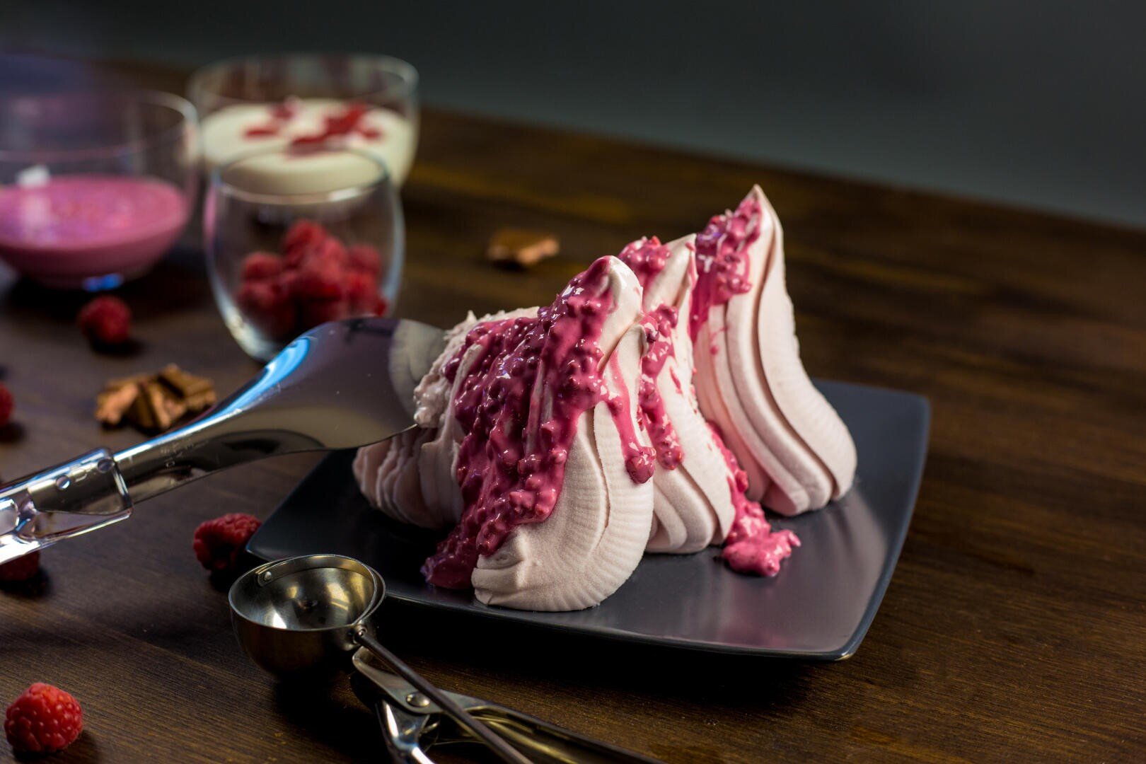 A dessert of piped, swirled pink meringue topped with raspberry sauce sits on a gray plate. Fresh raspberries, metal scoops, and glasses of creamy desserts are on a dark wooden table in the background.