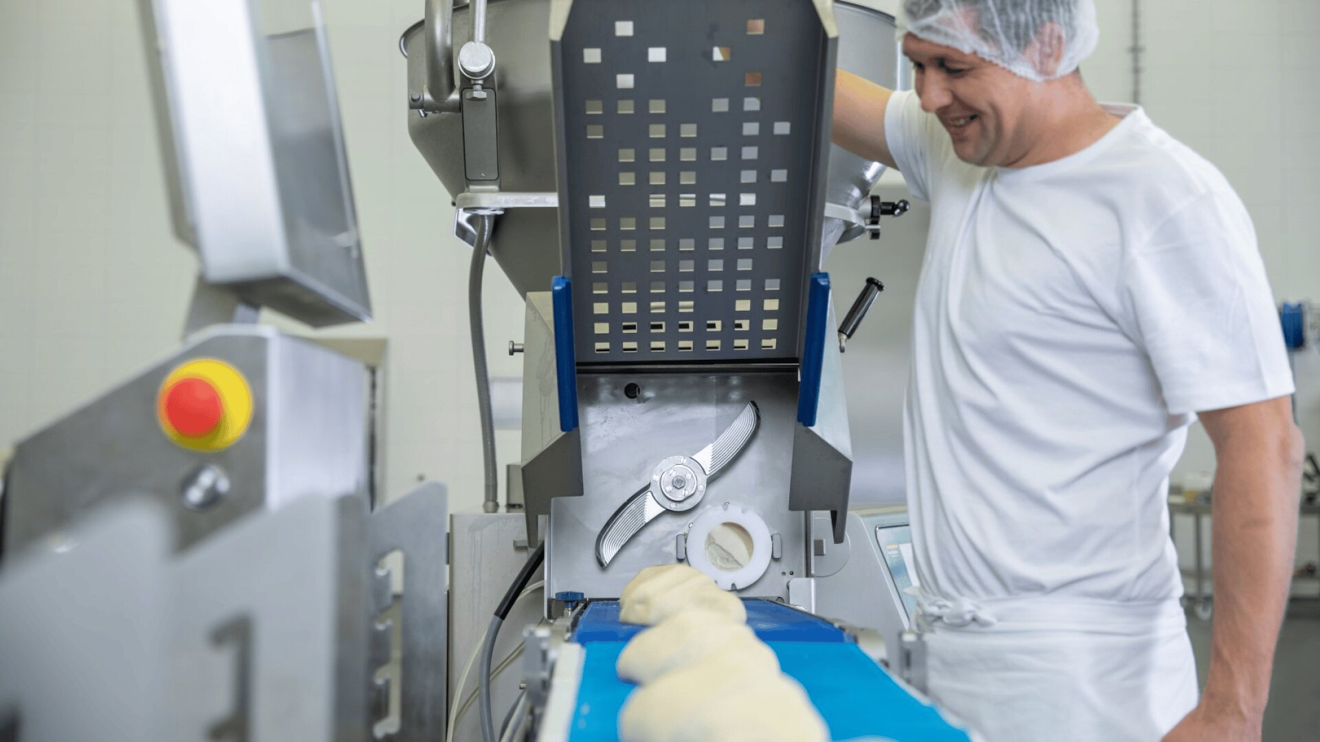 Ein Mann in weißer Uniform und mit Haarnetz lächelt, während er eine industrielle Teigverarbeitungsmaschine bedient, bei der sich Teigstücke über ein blaues Förderband bewegen.