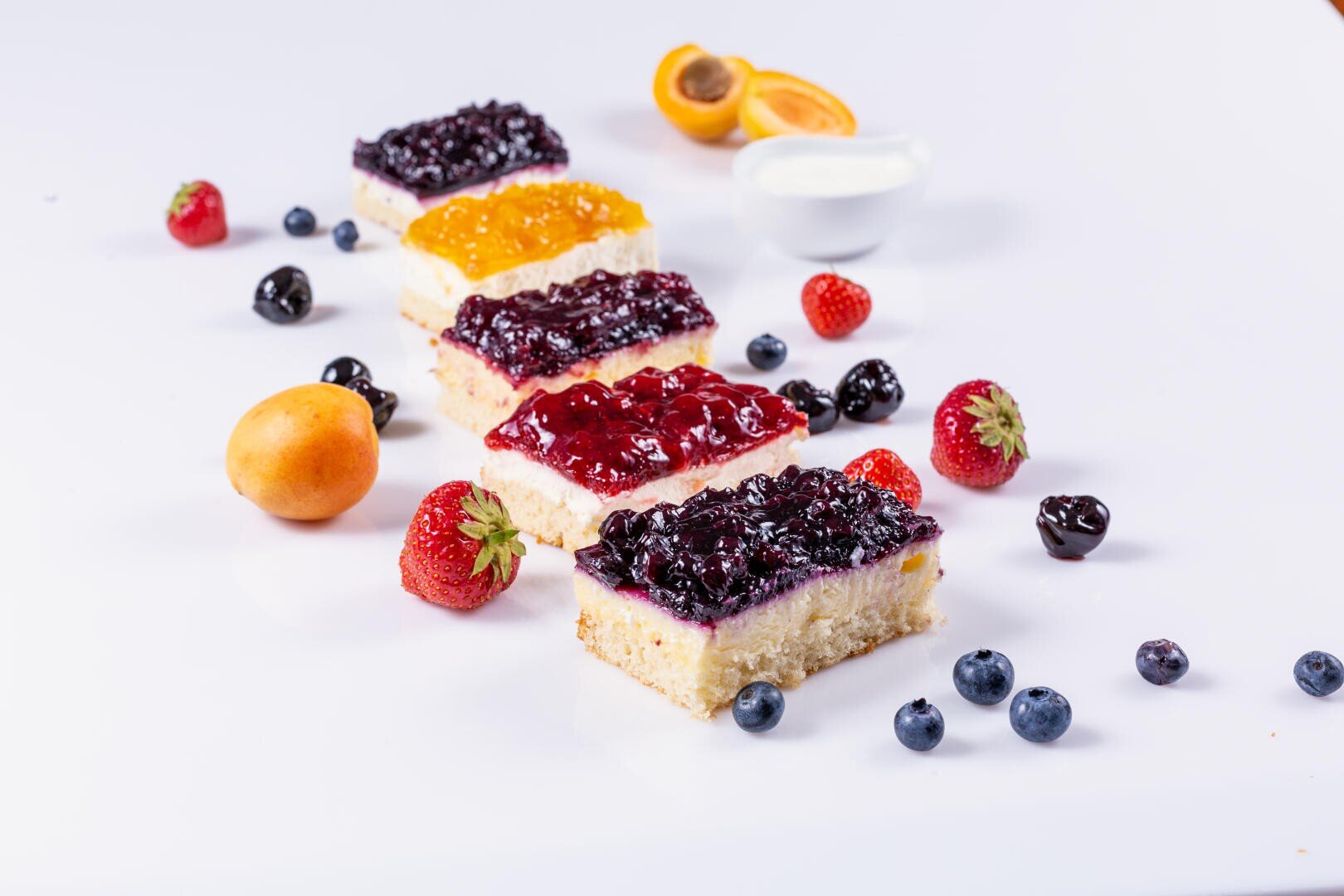Five rectangular fruit-topped cheesecake bars are arranged diagonally on a white surface, surrounded by fresh strawberries, apricots, blueberries, and a small bowl of cream.