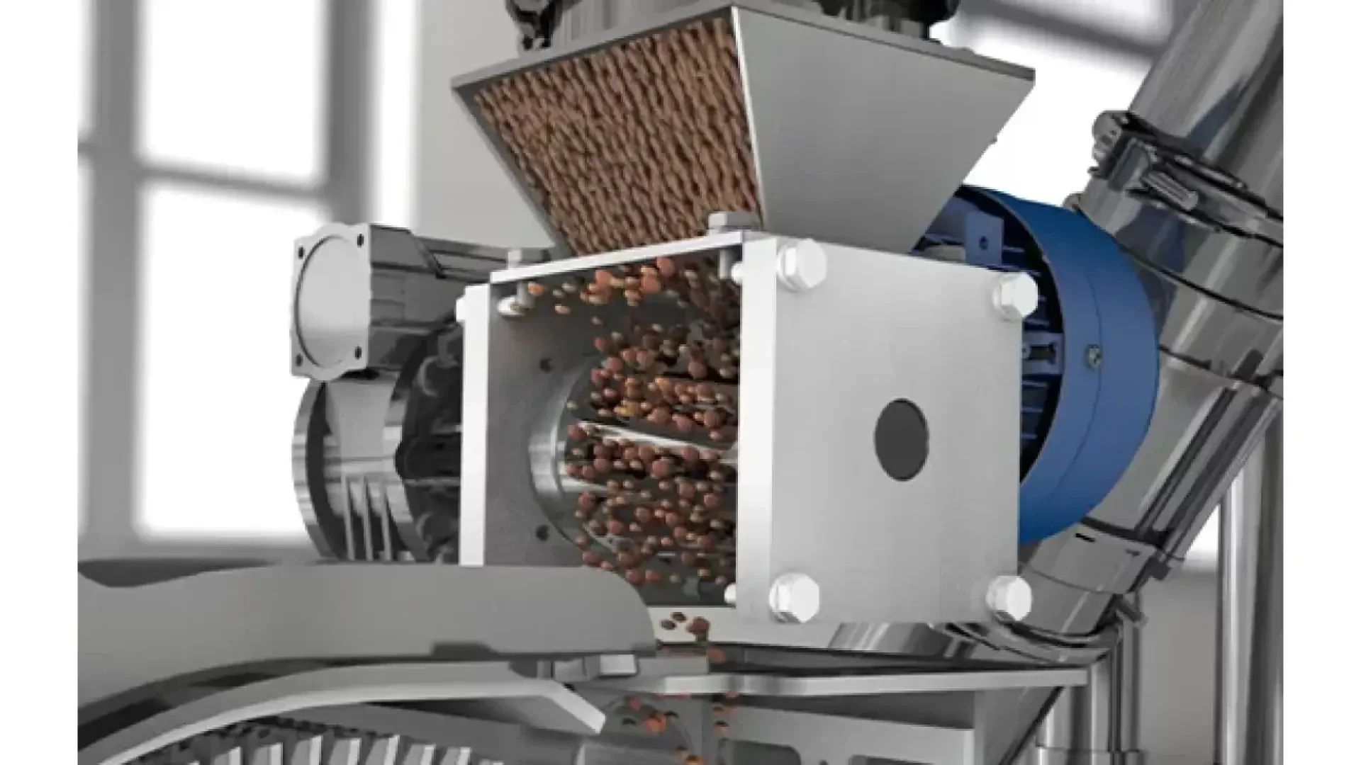 A close-up view of a food processing machine with brown pellets entering a metal chamber, possibly for grinding or mixing, in an industrial setting.
