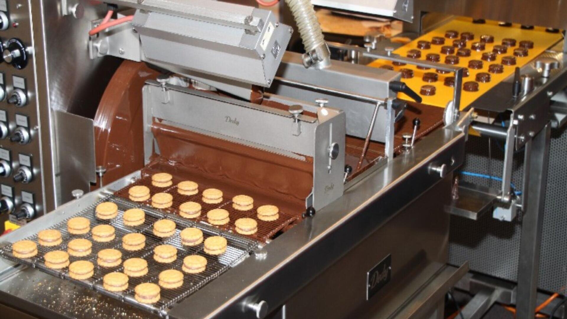 Cookies are traveling on a conveyor belt through a machine that is coating them with chocolate in a food processing facility. Rows of cookies are visible both before and after being coated.