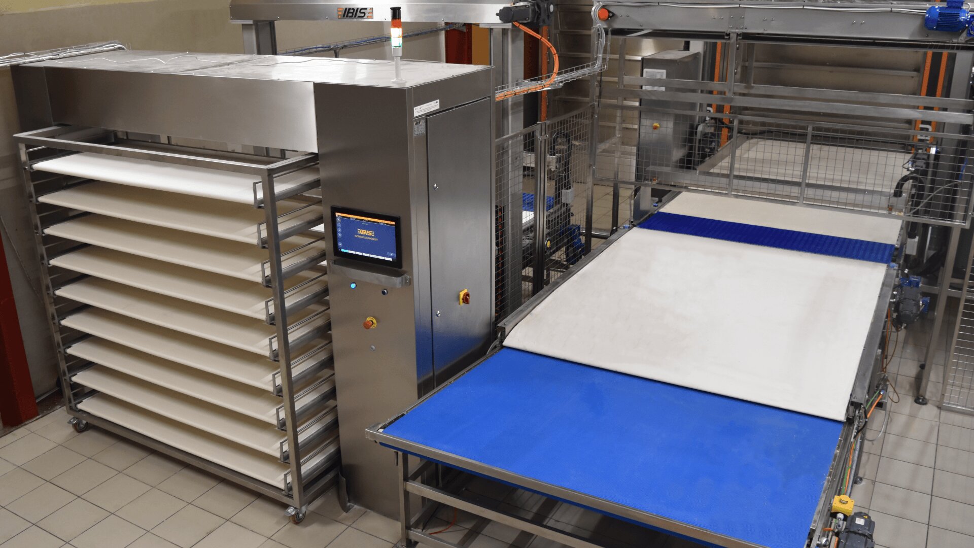 A modern industrial bakery machine with multiple metal racks, a digital control panel, and conveyor belts for processing dough, set in a tiled factory environment.