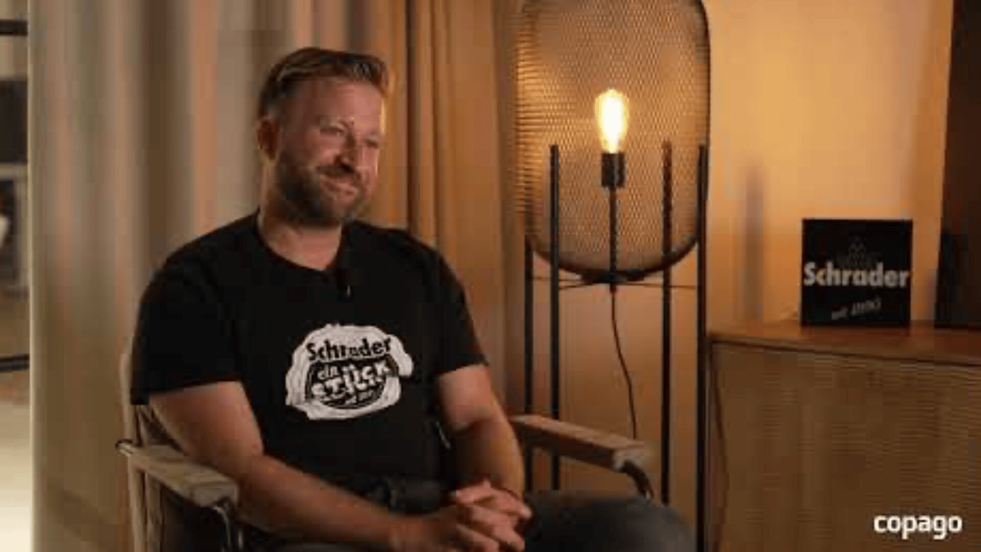 Ein bärtiger Mann sitzt lächelnd auf einem Stuhl und trägt ein schwarzes "Schrader und Stick"-T-Shirt. Eine beleuchtete Stehlampe und ein Schrader und Stick-Schild befinden sich im Hintergrund. Die Szene hat eine warme, gemütliche Atmosphäre.