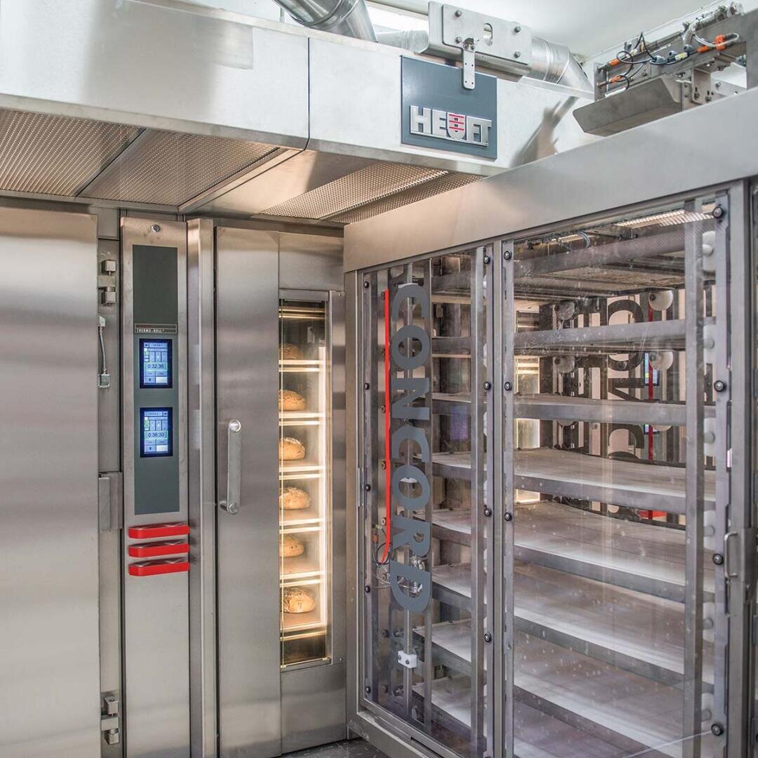 Ein moderner industrieller Backofen mit mehreren Backblechen im Inneren, mit digitalen Bedienfeldern und Edelstahloberflächen in einer sauberen, gut beleuchteten Umgebung. Der Markenname HEUFT ist auf der Maschine sichtbar.