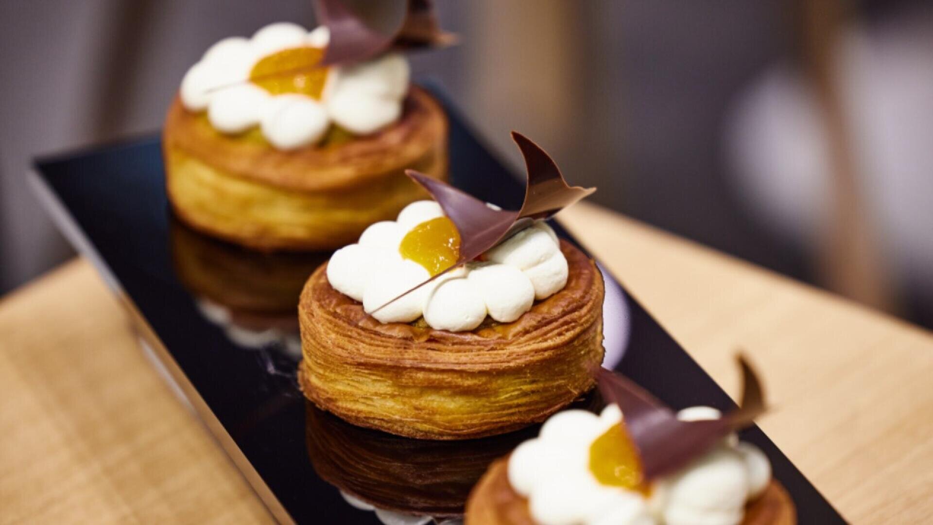 Three round pastries topped with white cream, a dollop of yellow jam, and decorative dark chocolate pieces are arranged on a glossy black tray set on a light wooden surface.