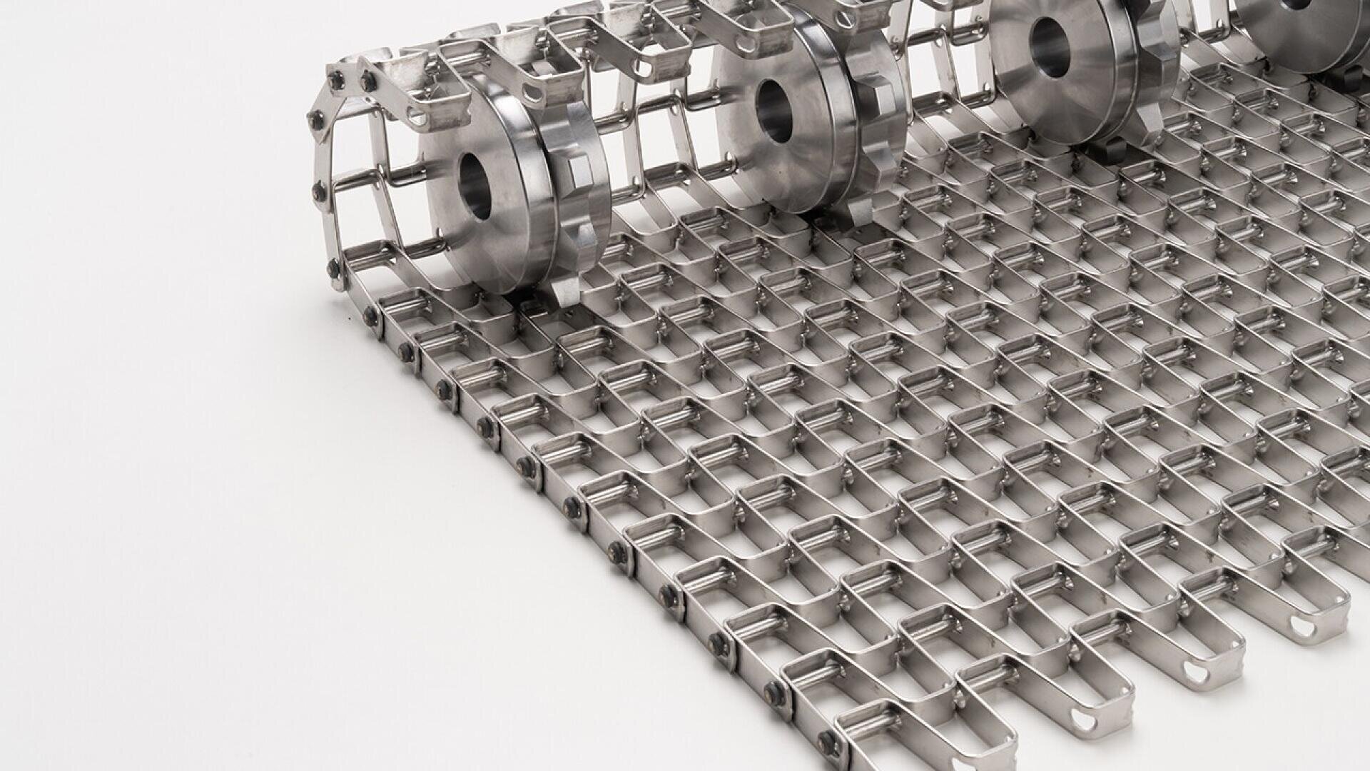 Nahaufnahme eines Förderbandes aus Edelstahl mit einem Metallgittermuster und zylindrischen Rollen an einem Ende, auf einem weißen Hintergrund positioniert.