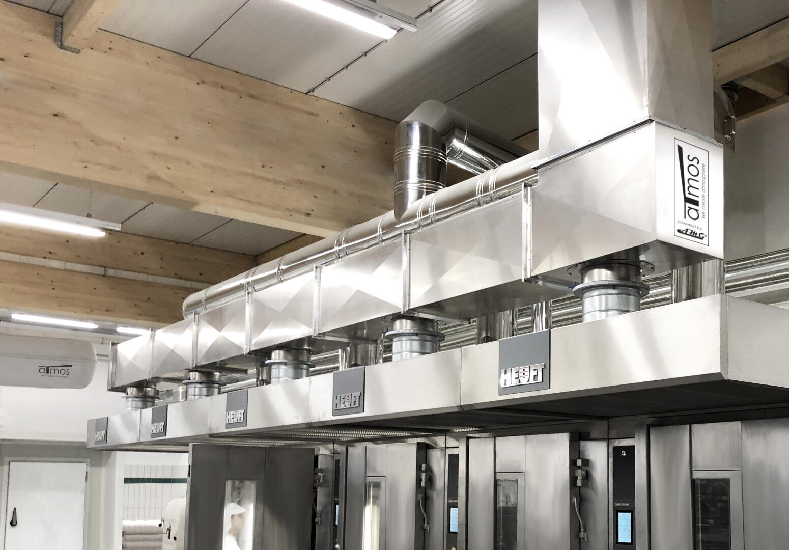 Large industrial stainless steel ventilation system installed above commercial ovens in a bakery or food production facility with wooden ceiling beams and bright lighting.