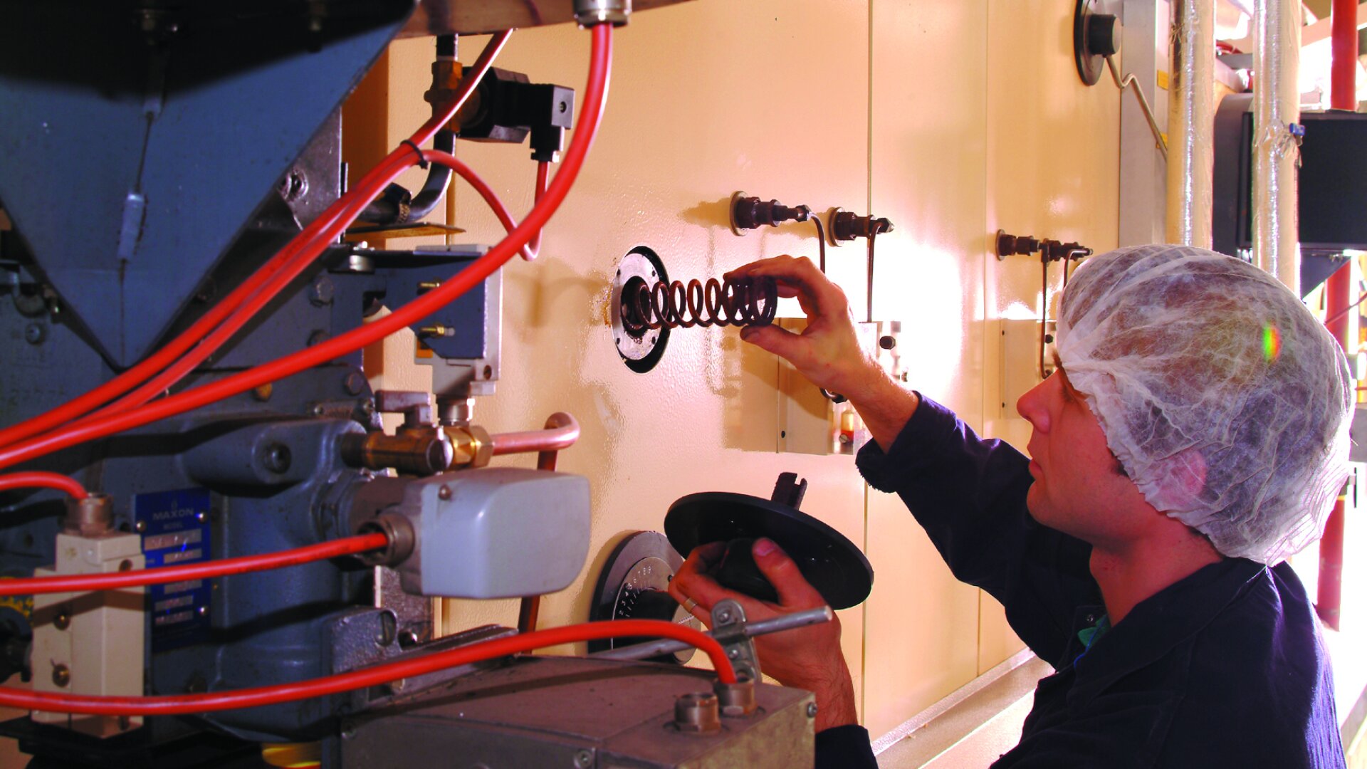 A worker wearing a hairnet adjusts a large machine panel, holding a circular metal spring and a cover, with red cables and industrial equipment visible.