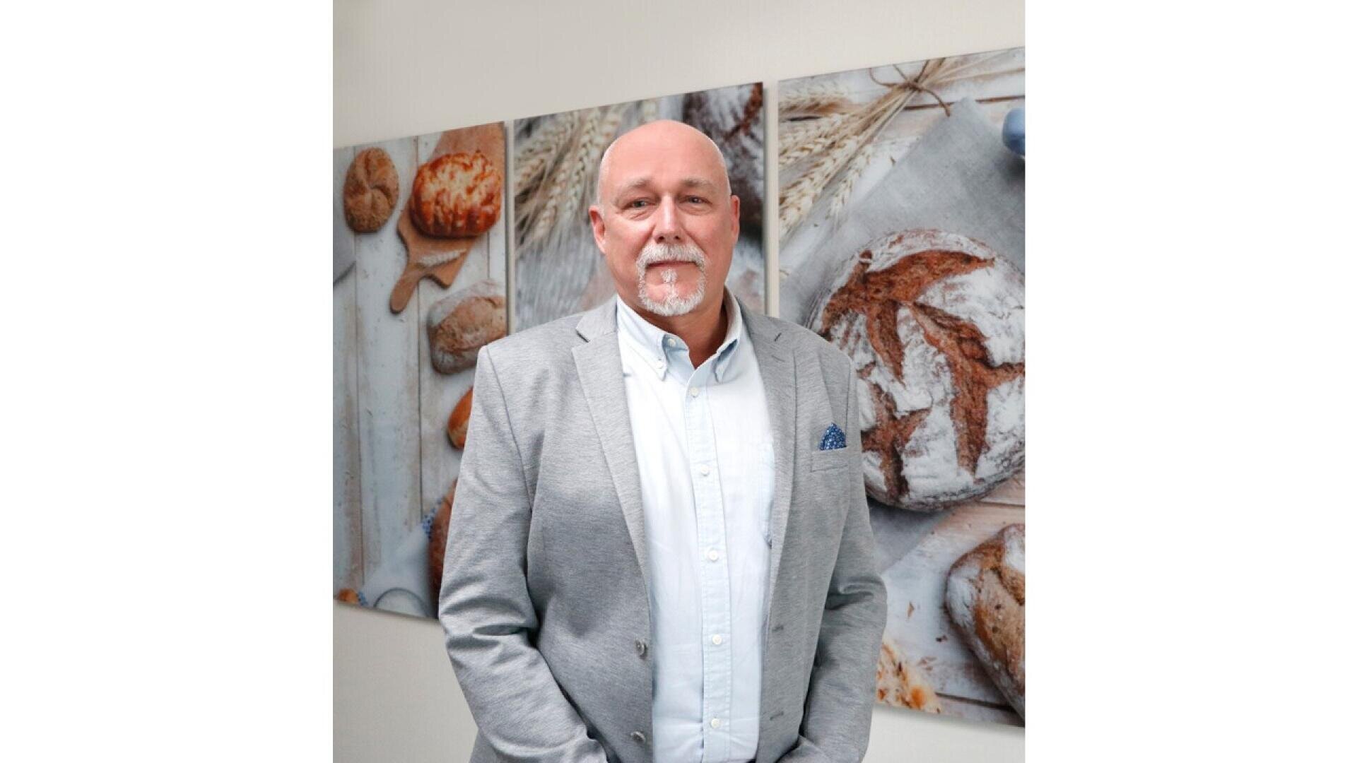 A man with a bald head and white beard, wearing a light gray blazer over a button-up shirt, stands in front of wall art featuring images of rustic bread and wheat.