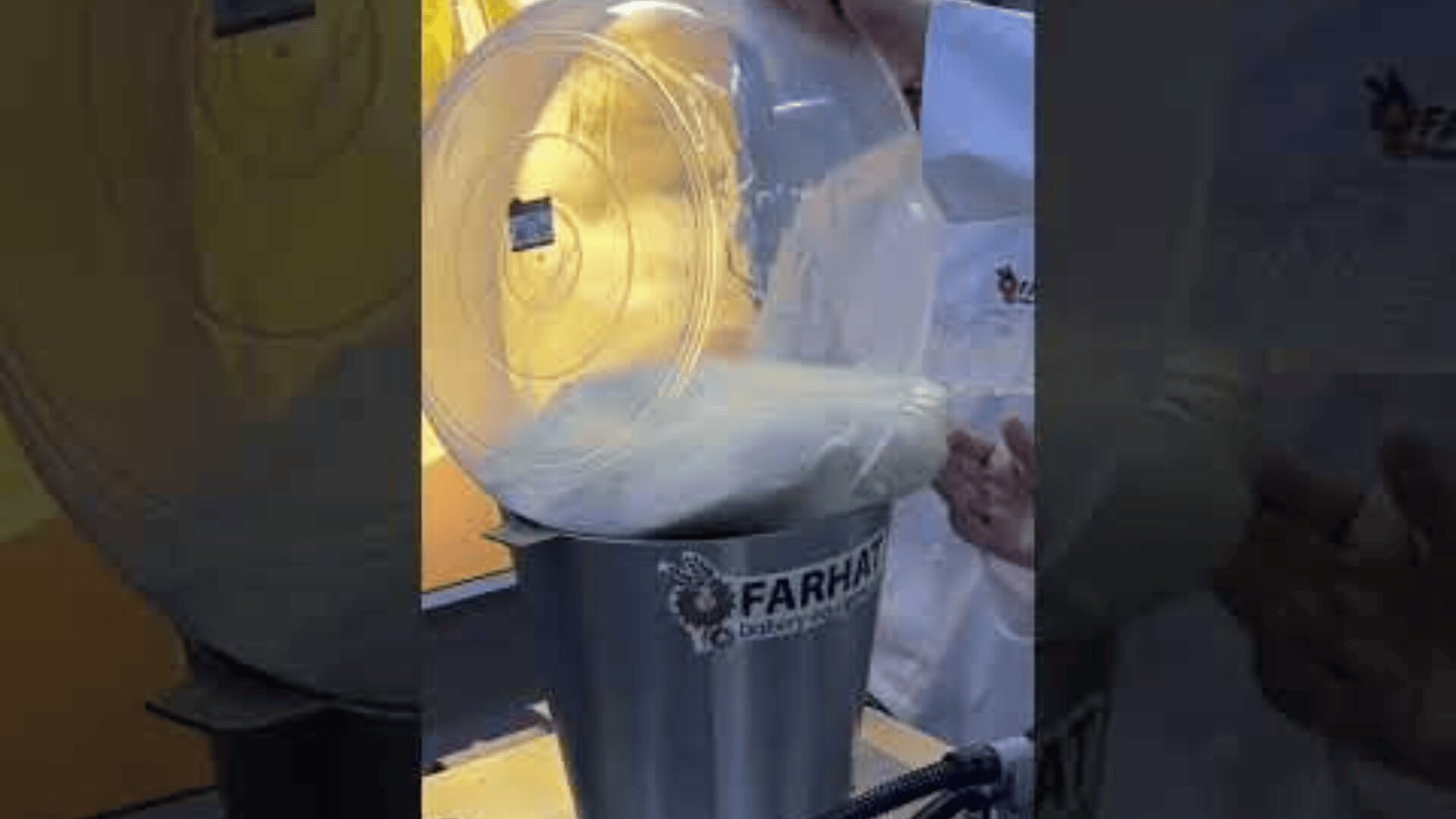 A person pours white flour from a large transparent container into a metal machine labeled FARHA, which appears to be in a commercial kitchen or bakery.