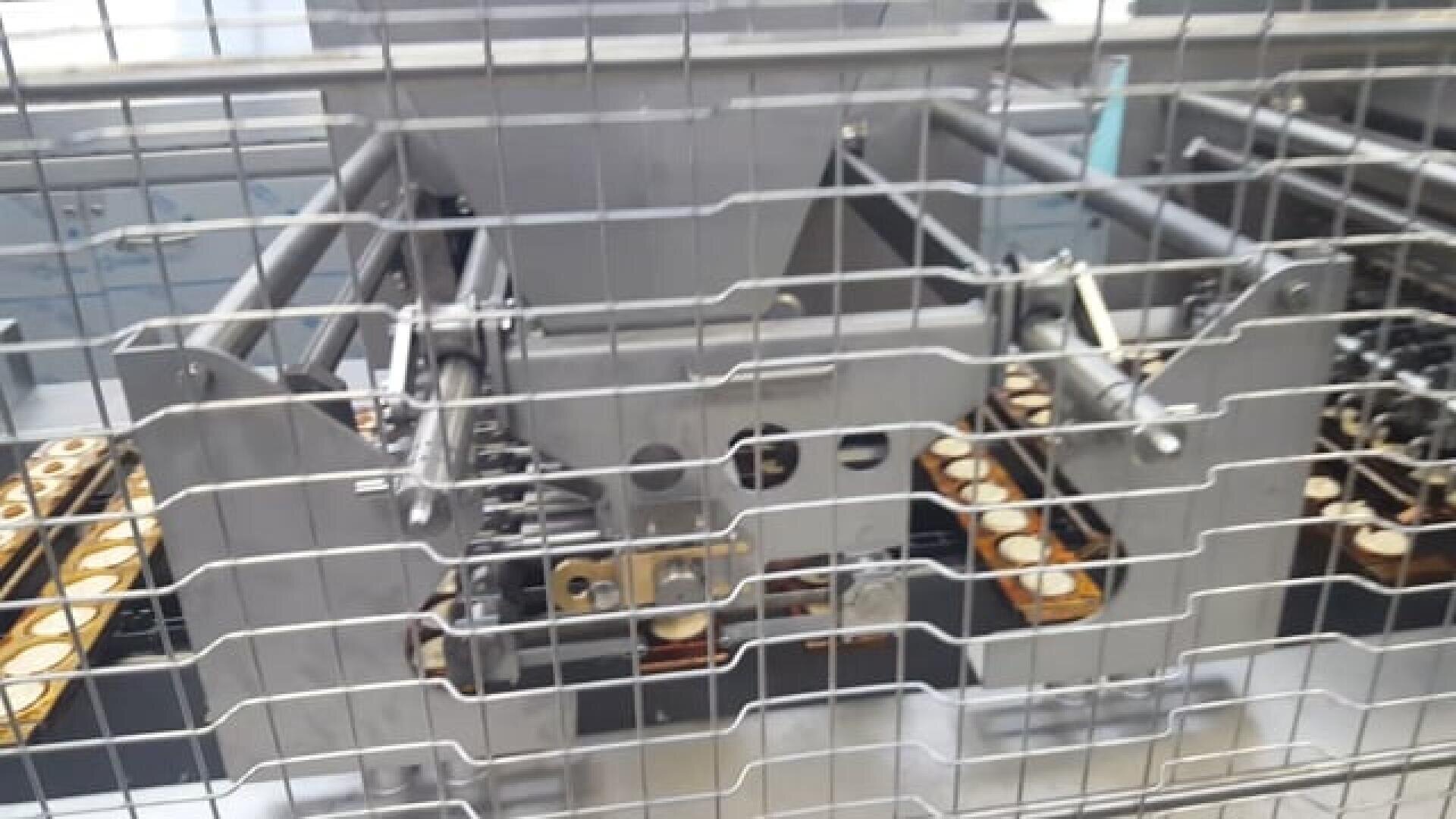 Close-up of an industrial baking machine behind a metal safety cage, with rows of small round pastry shells arranged on trays inside the machine.