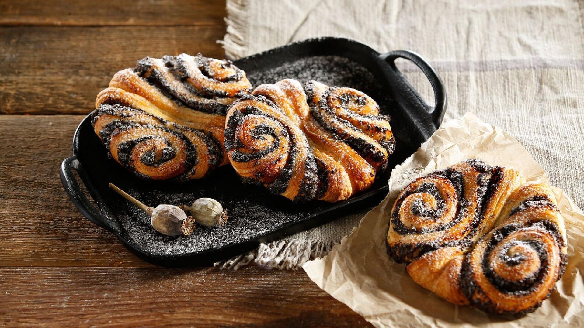 Three swirled chocolate pastries dusted with powdered sugar are displayed on a black tray and parchment paper atop a rustic wooden table and a beige cloth. Two dried poppy pods are also placed on the tray.