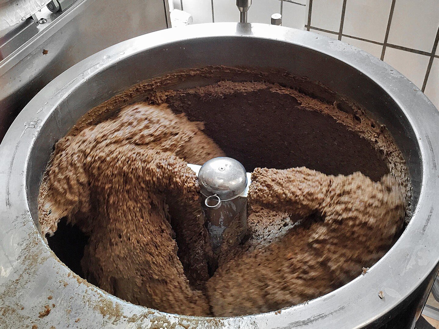 Ein großer industrieller Mixer mischt eine dicke, braune Mischung in einem Metallbehälter, wobei der Inhalt um das zentrale Mischblatt herumgewirbelt wird. Im Hintergrund sind weiß gekachelte Wände zu sehen.