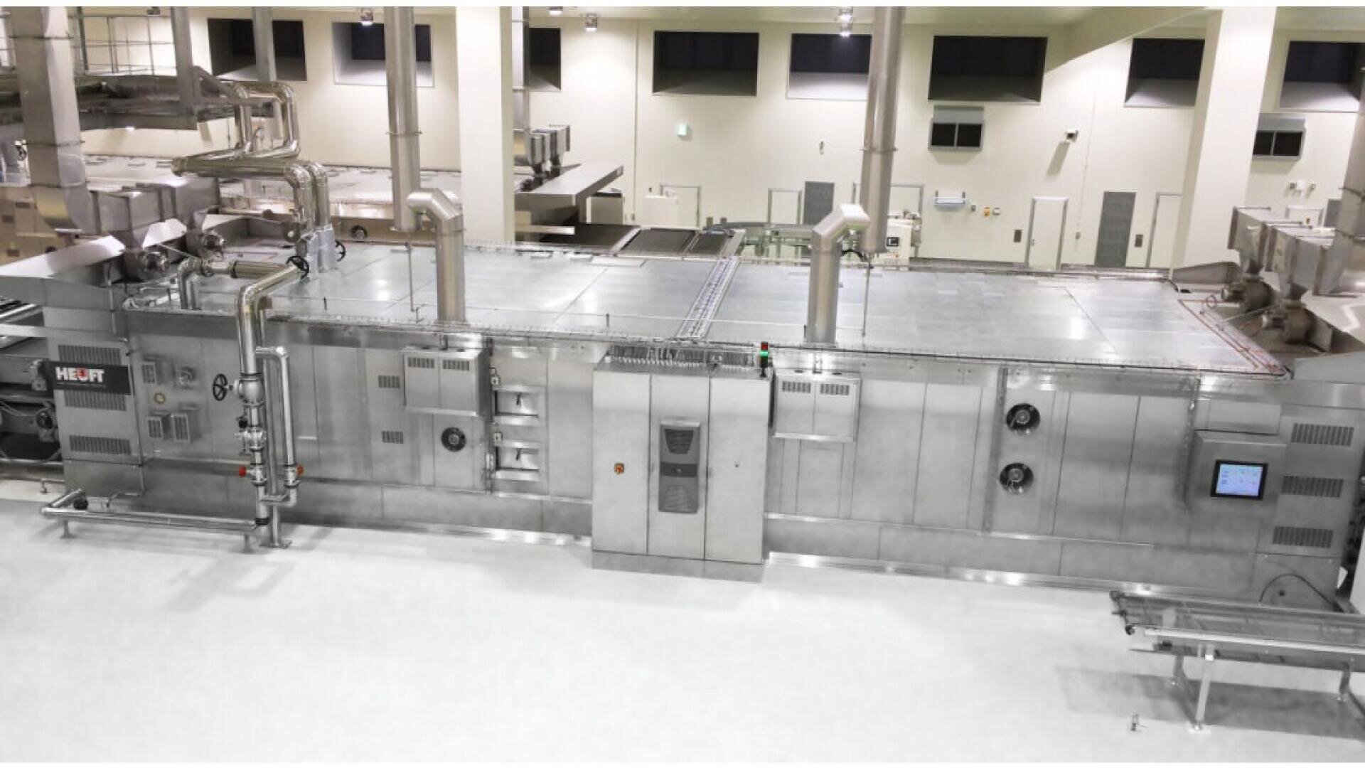 A large, modern industrial food processing machine with stainless steel panels, pipes, and control screens in a clean, well-lit factory setting. The floor is white and there are no workers visible.