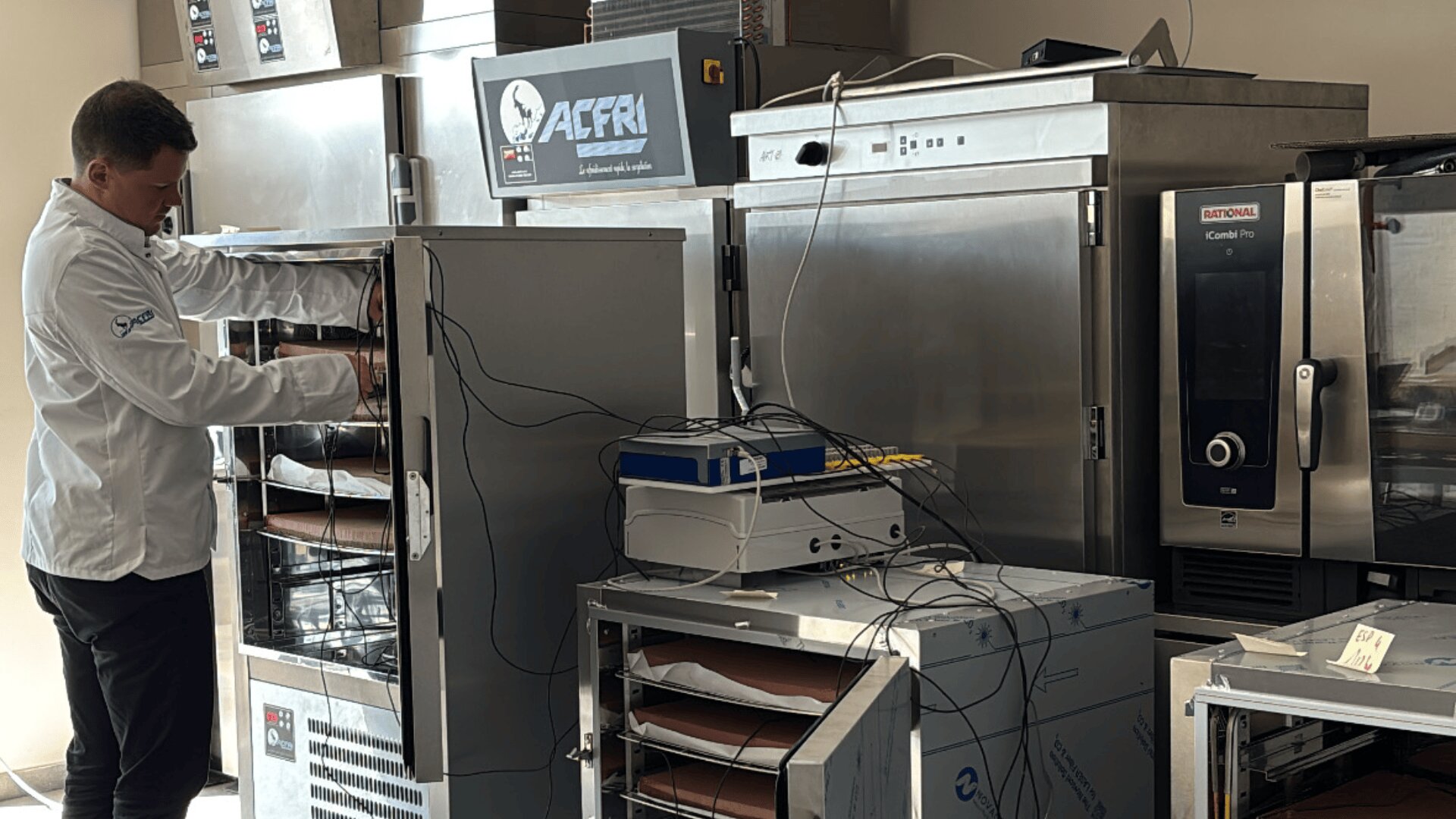 Ein Mann im weißen Laborkittel justiert Geräte in einer Großküche mit mehreren Edelstahlöfen und -geräten. Kabel verbinden die verschiedenen Geräte, und Sonnenlicht fällt durch ein nahes Fenster.
