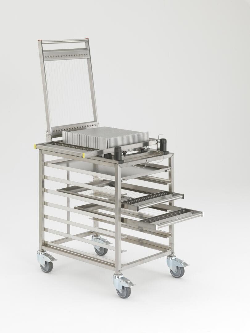 Ein medizinischer oder labortechnischer Wagen aus Edelstahl mit Rädern, mehreren offenen, verschiebbaren Fächern und einem aufrecht stehenden Gerät auf der Oberseite, das eine Schneidemaschine zu sein scheint, vor einem schlichten weißen Hintergrund.