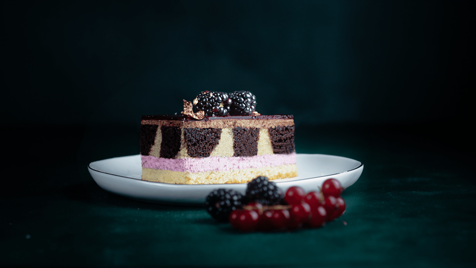 A slice of layered cake with chocolate, vanilla, and pink cream on a white plate, garnished with blackberries and a gold decoration, with blackberries and redcurrants beside the plate on a dark background.