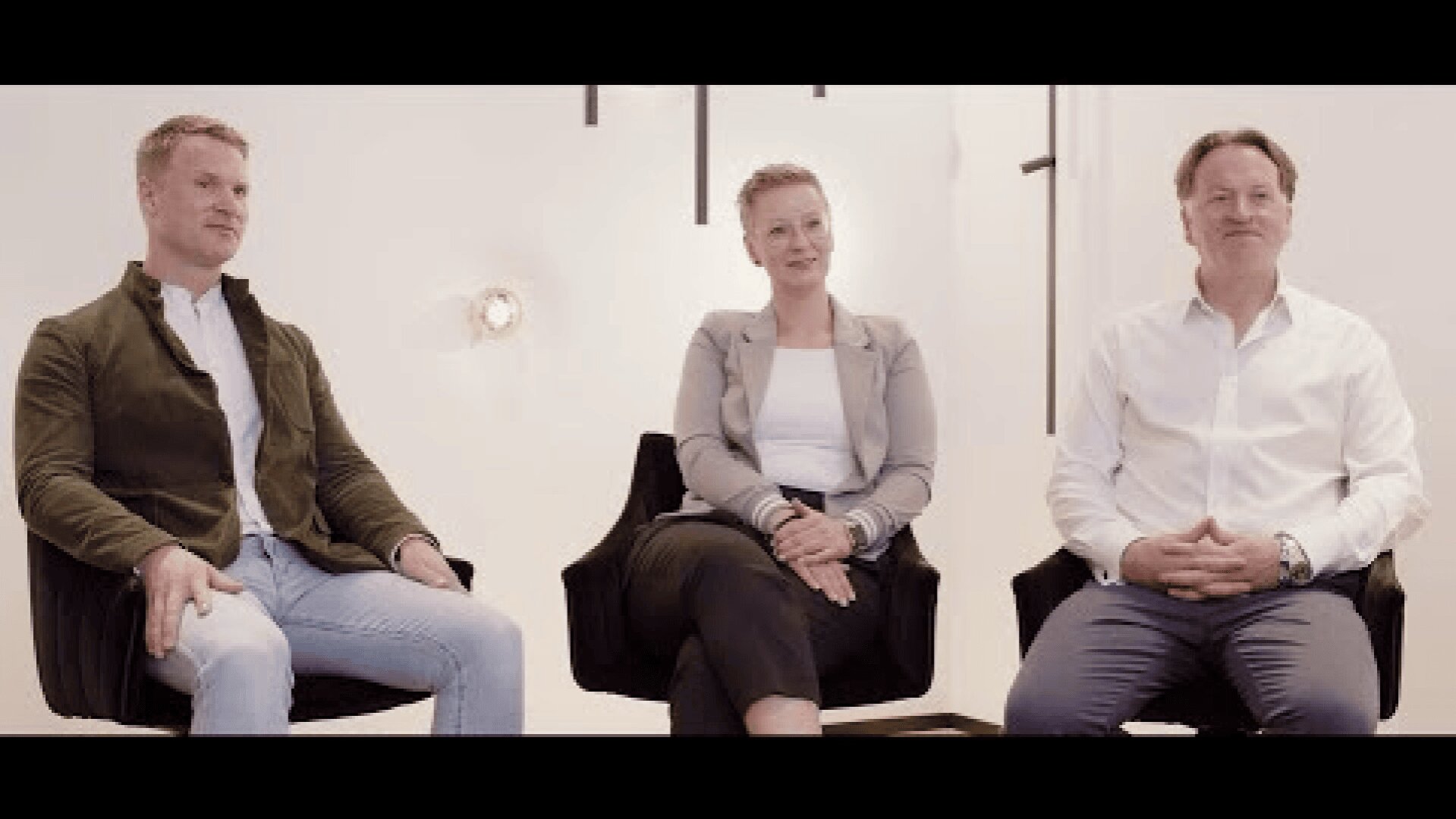 Drei Personen sitzen auf schwarzen Stühlen vor weißem Hintergrund. Zwei Männer, einer in grüner Jacke und Jeans, der andere in weißem Hemd, sitzen zu beiden Seiten einer Frau in hellem Blazer und schwarzem Rock. Sie blicken in die Kamera und scheinen sich zu unterhalten.