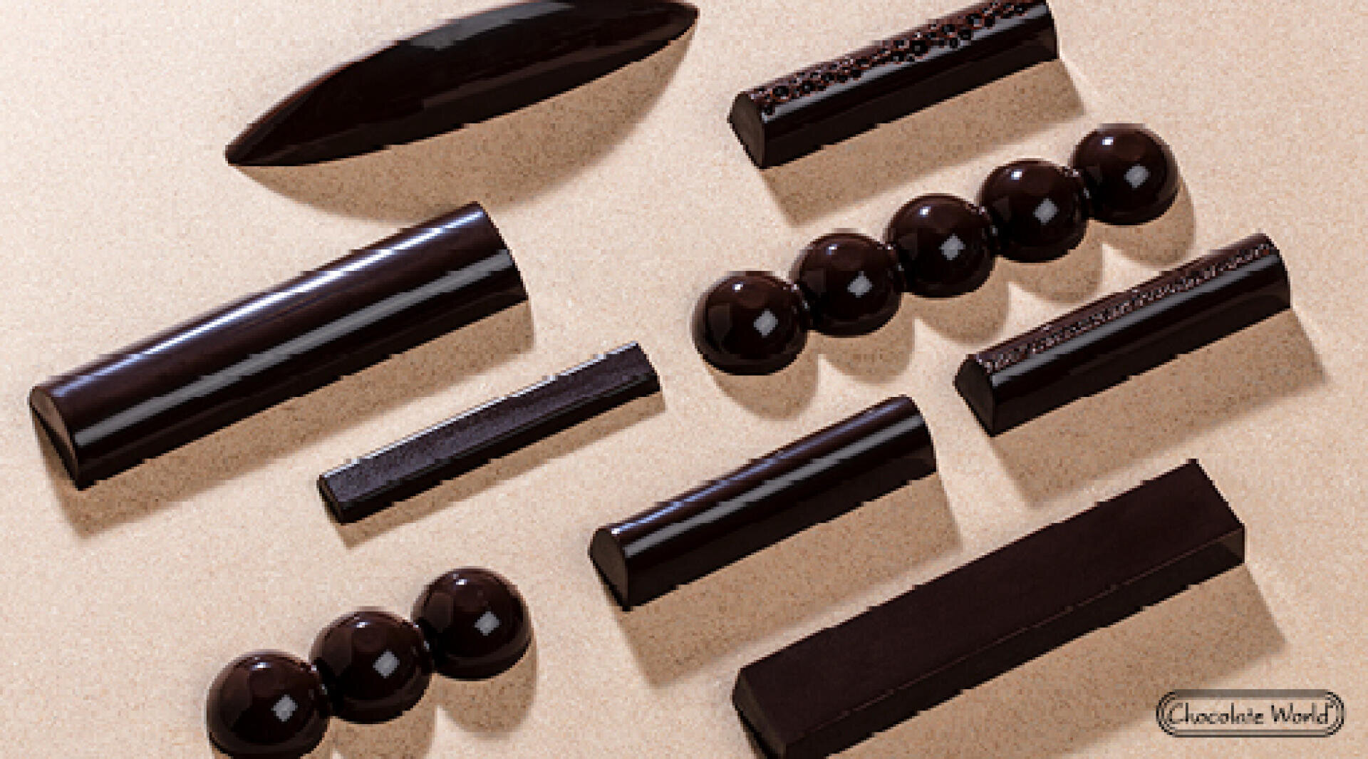 Several glossy, dark chocolate pieces in various geometric shapes, including spheres, bars, and domes, are arranged neatly on a light beige surface with a Chocolate World logo in the corner.