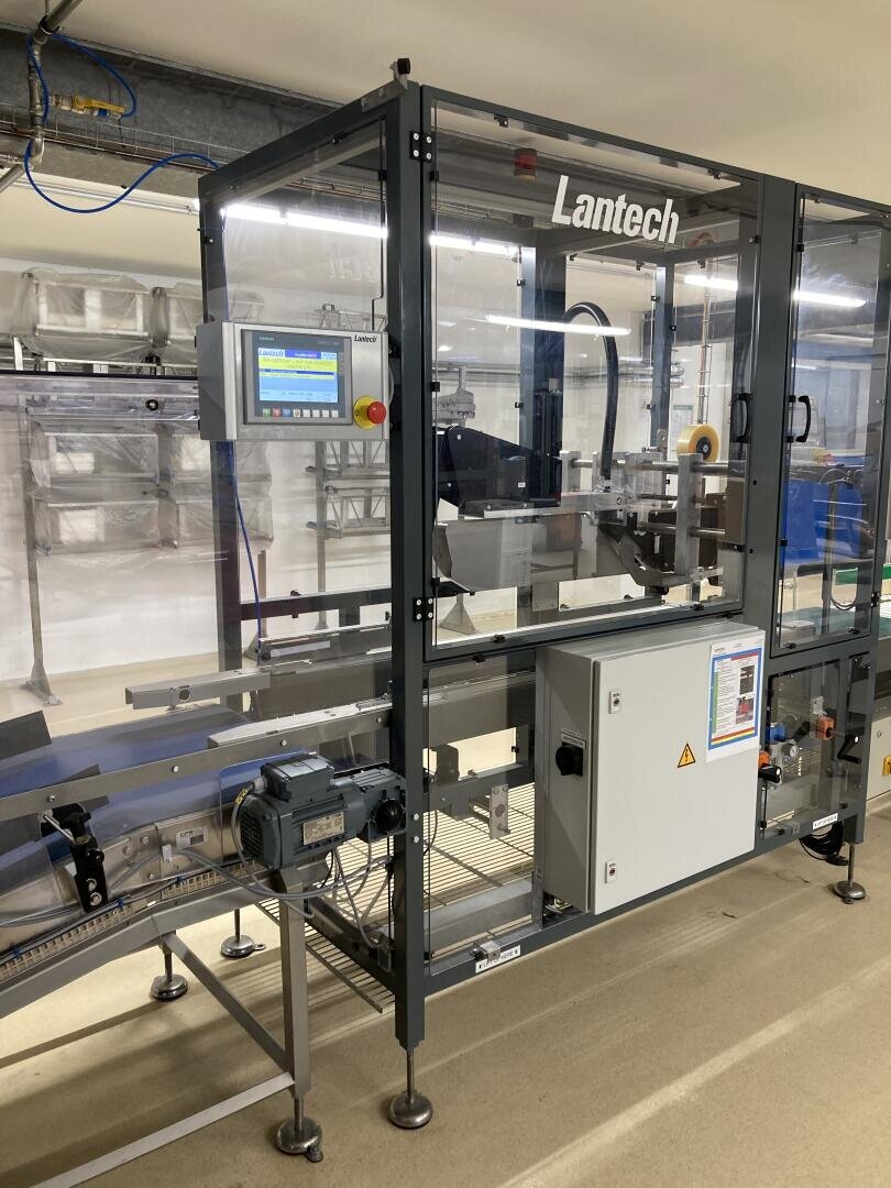 A Lantech automated packaging machine with a touch screen control panel stands in a clean industrial facility, surrounded by metal conveyor belts and electrical boxes.