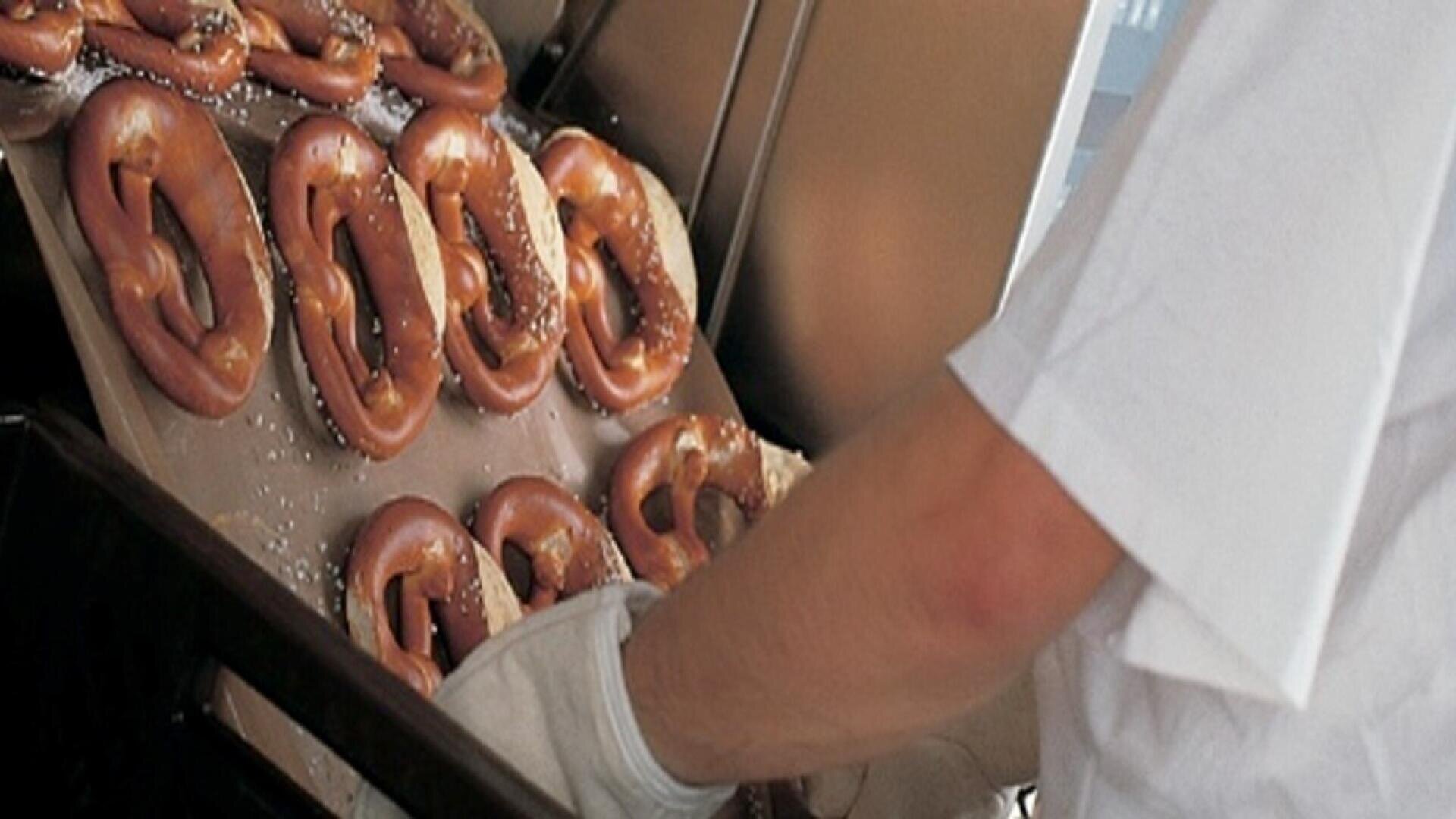 A person wearing a white shirt and glove removes a tray of freshly baked pretzels from an oven; the pretzels are golden-brown and sprinkled with coarse salt.
