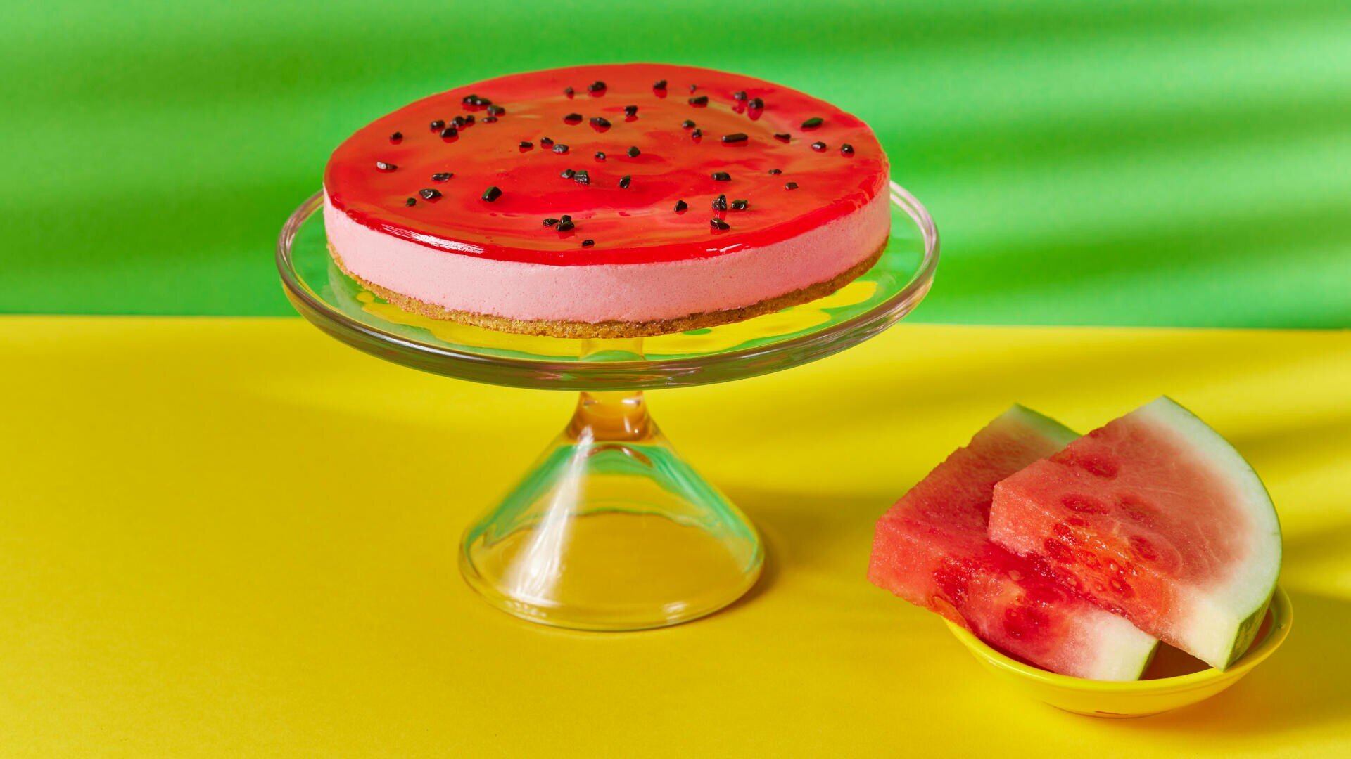 Ein von Wassermelonen inspirierter Käsekuchen mit einer rosa Schicht und einer glänzend roten Oberseite, garniert mit Schokoladenstückchen, die an Kerne erinnern, steht auf einem Glasständer neben Wassermelonenscheiben auf einem gelben und grünen Hintergrund.