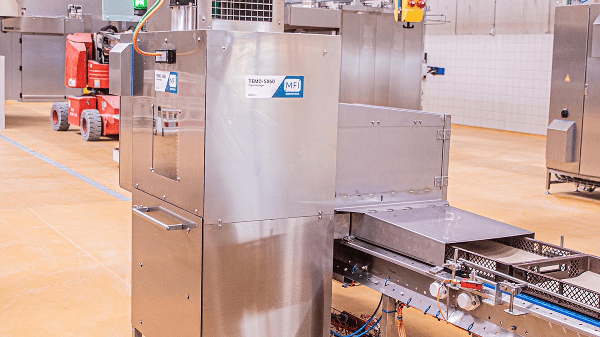 A stainless steel industrial food processing machine stands in a factory setting, with various control panels and compartments visible. Conveyor belts and other machinery appear in the background on a polished floor.