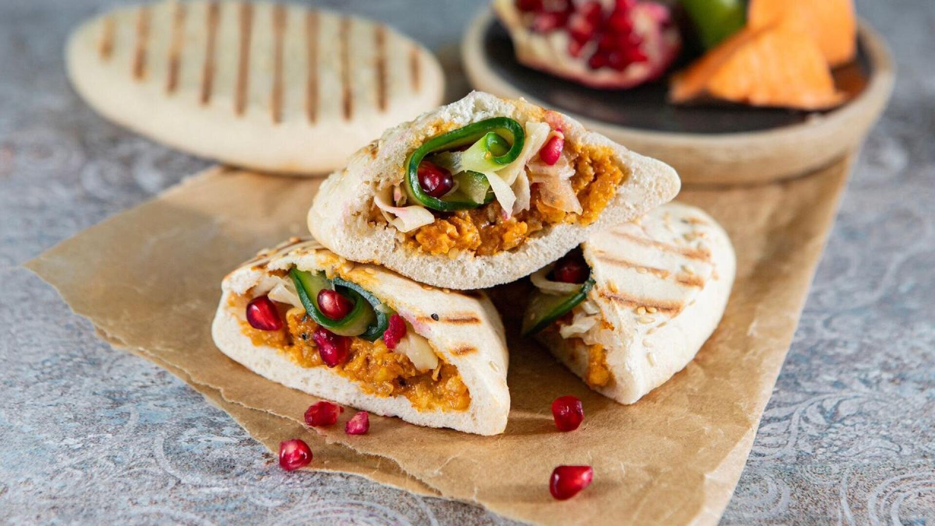 Drei gegrillte Pita-Taschen gefüllt mit Orangenaufstrich, Hühnerstreifen, Gurkenscheiben und Granatapfelkernen, auf Backpapier gestapelt, mit zusätzlichen Pitas und Obst im Hintergrund.