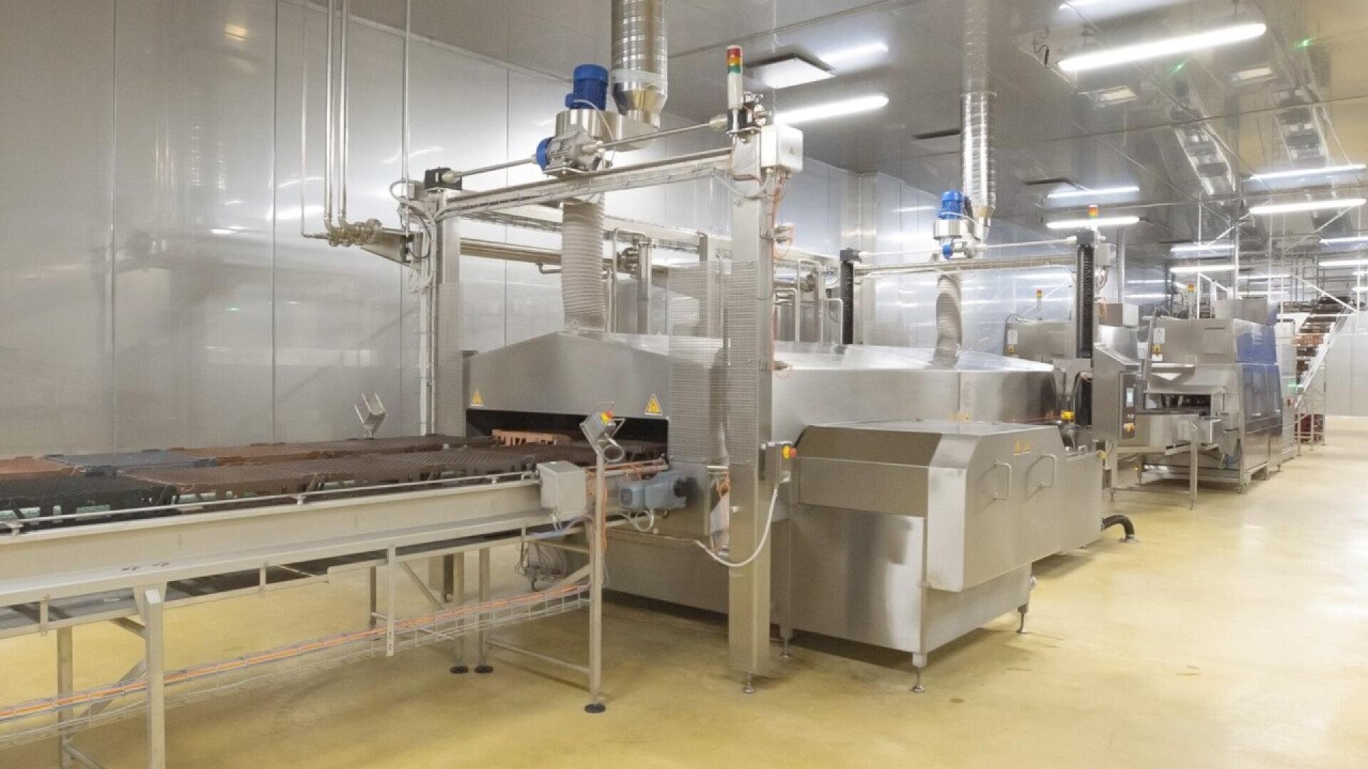 A modern industrial food processing machine in a clean, brightly lit factory setting, with conveyor belts and stainless steel equipment visible.