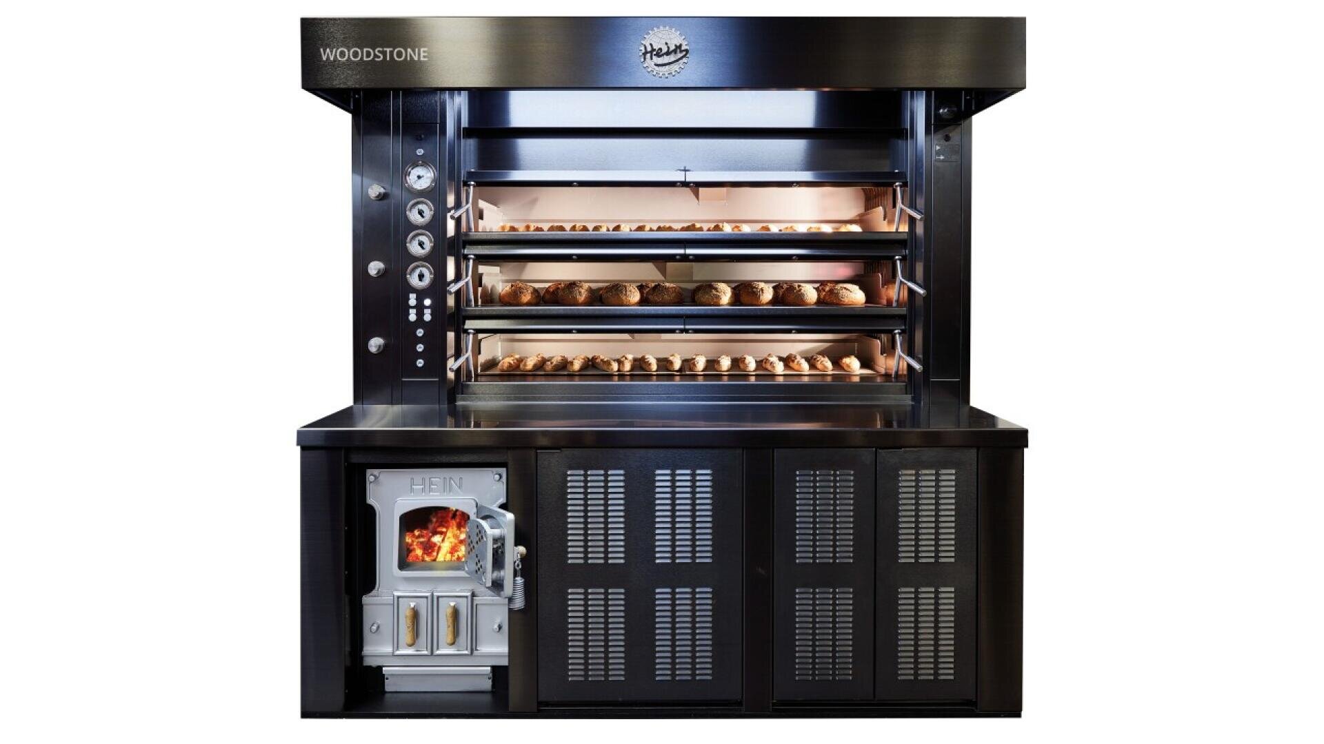 A commercial bread oven with three shelves full of loaves baking, metal controls on the left, and a wood-fired compartment with a visible fire at the bottom left. The oven is labeled WOODSTONE and Heidis.