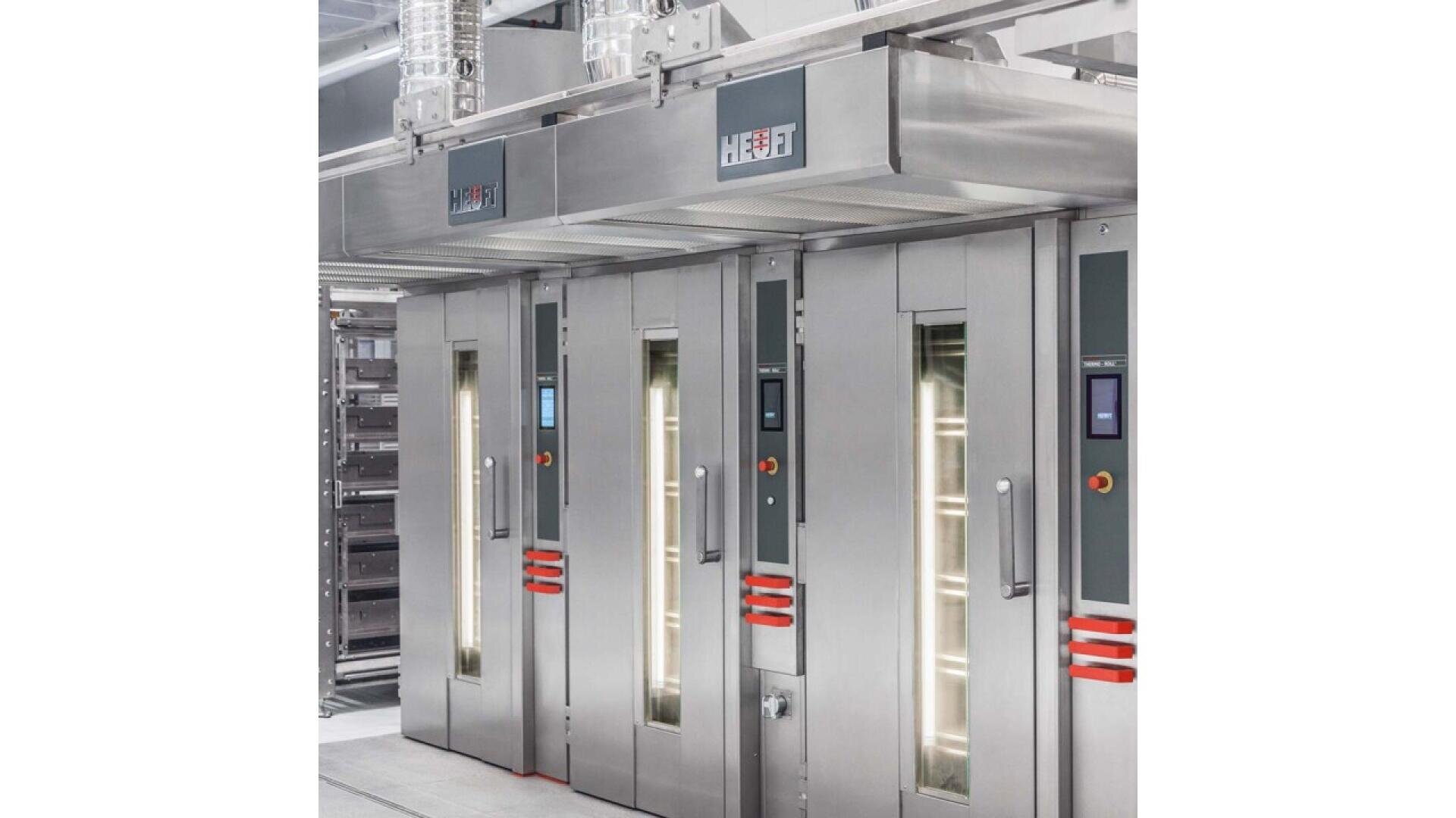 A row of large, stainless steel industrial ovens with glass doors and control panels, inside a modern commercial kitchen or bakery.
