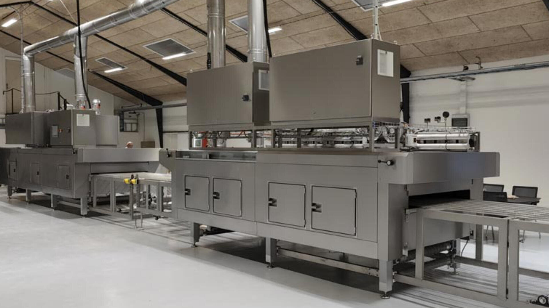 Große industrielle Lebensmittelverarbeitungsmaschine mit Edelstahlkomponenten, Förderbändern und Lüftungsrohren in einer sauberen, geräumigen Fabrikumgebung mit hellen Wänden und Deckenplatten.