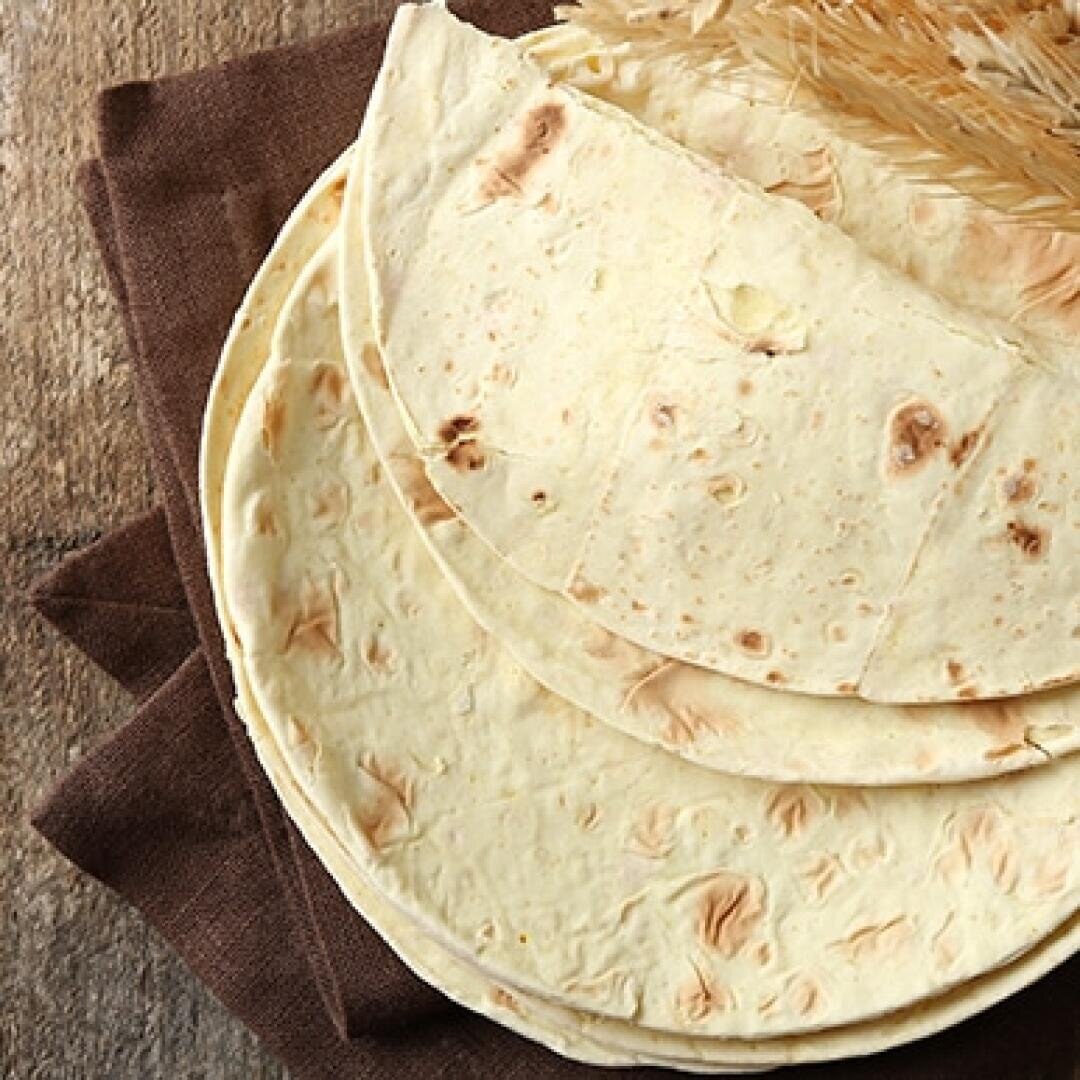 Four pieces of flatbread are stacked on a brown cloth napkin, with sprigs of wheat placed on top, all set on a rustic wooden surface.