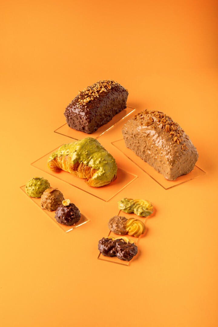 Two loaves of bread and two trays of assorted flavored cookies or pastries are arranged on a bright orange background. The pastries include green, brown, and yellow varieties.