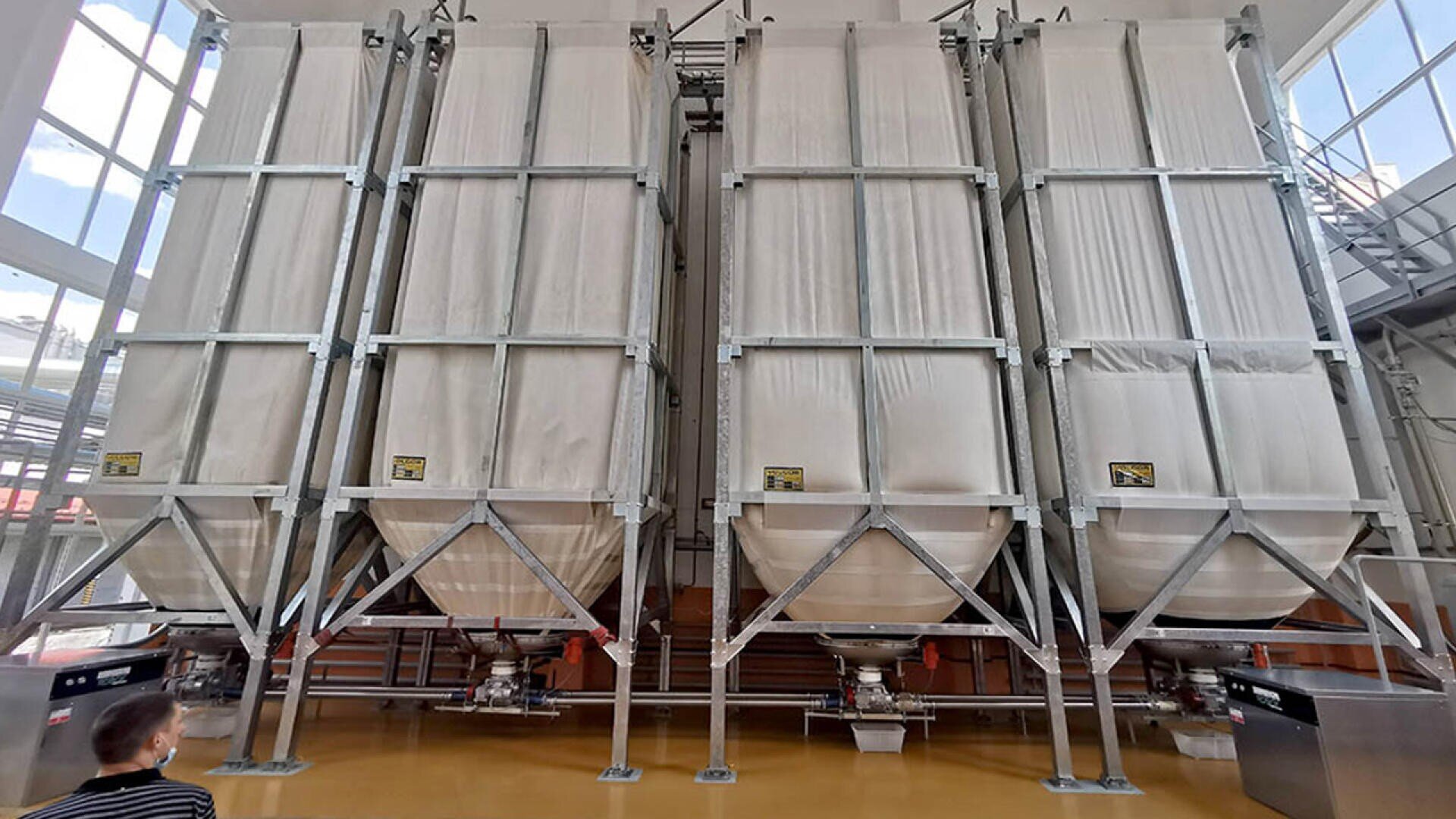 Large industrial fabric silos stand in a row inside a bright facility, supported by metal frames and containing bulk materials; a person is visible in the lower left corner for scale.
