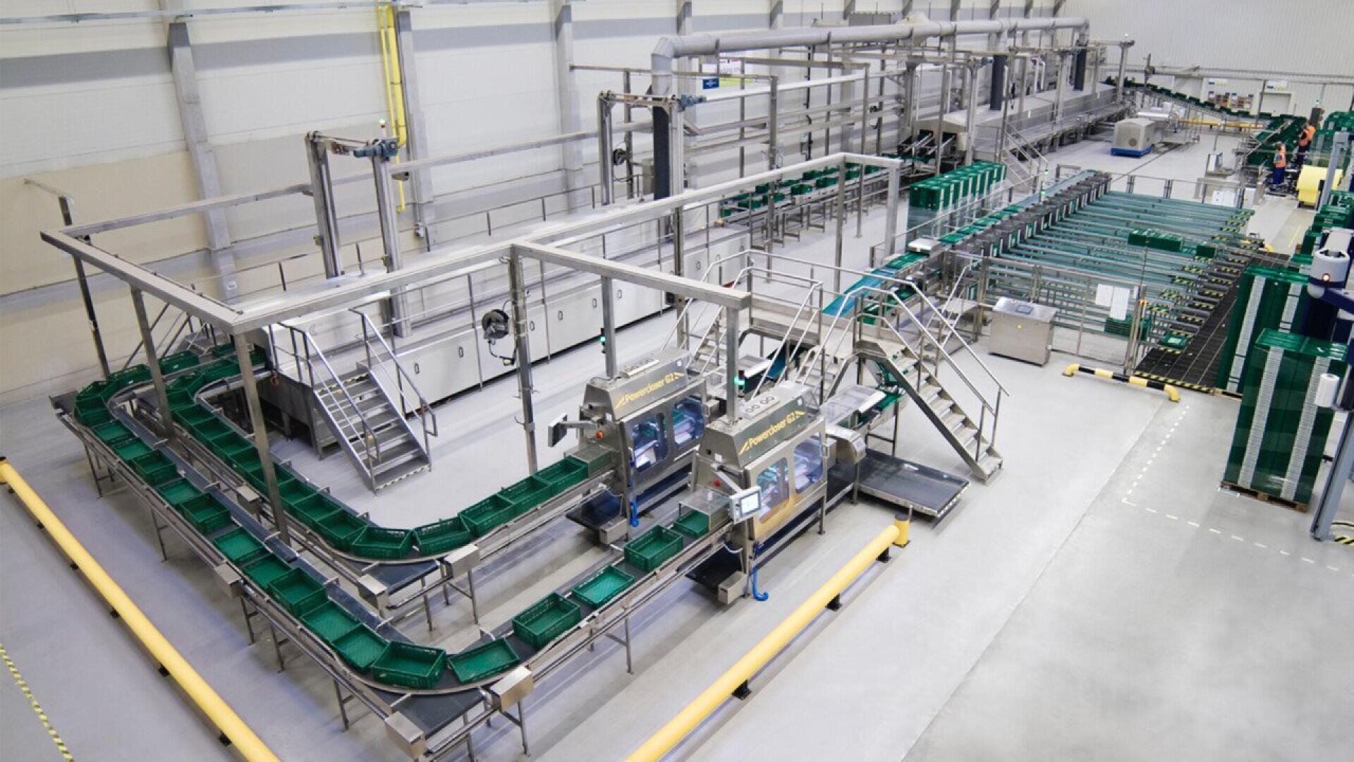 A modern factory interior showing an automated conveyor system with green trays, metal structures, and various machinery used for material handling and packaging. The area is clean, spacious, and well-lit.