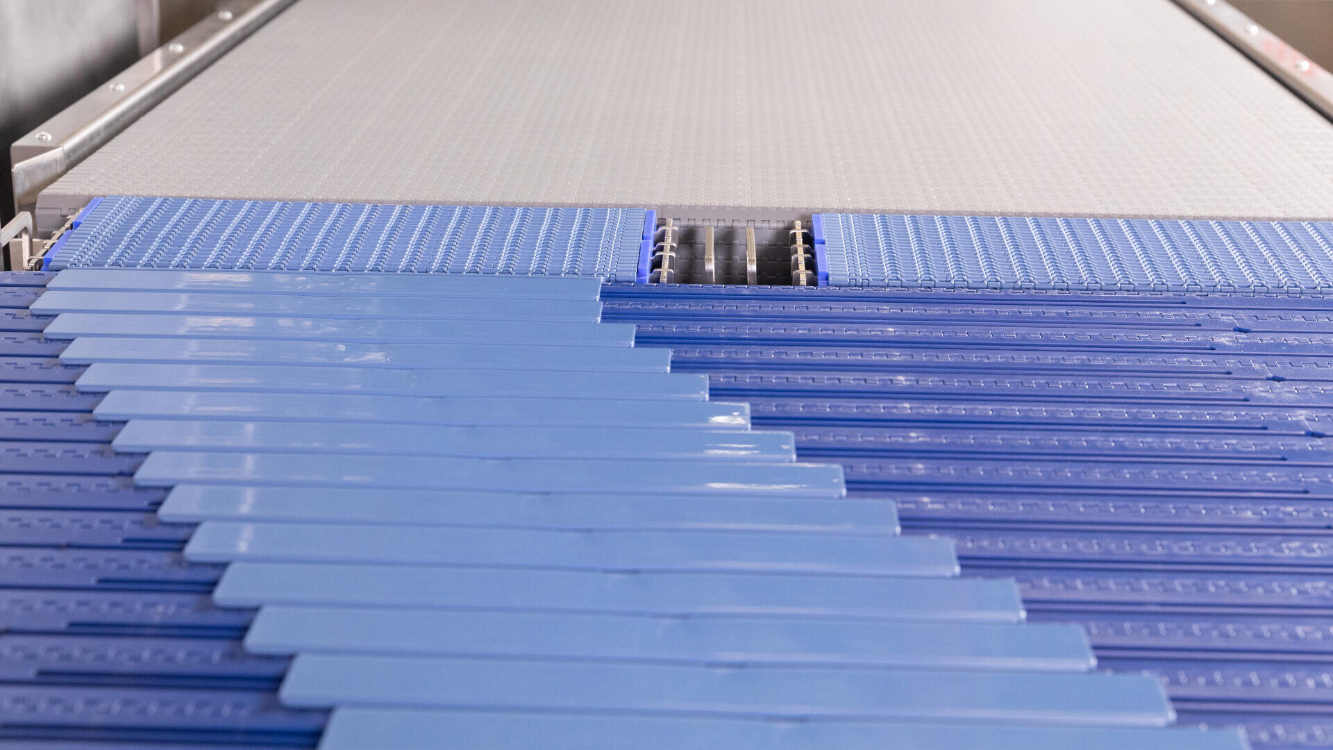 Close-up view of a blue conveyor belt system in a factory or industrial setting, with interlocking plastic slats arranged diagonally, transitioning onto a flat grey conveyor surface.