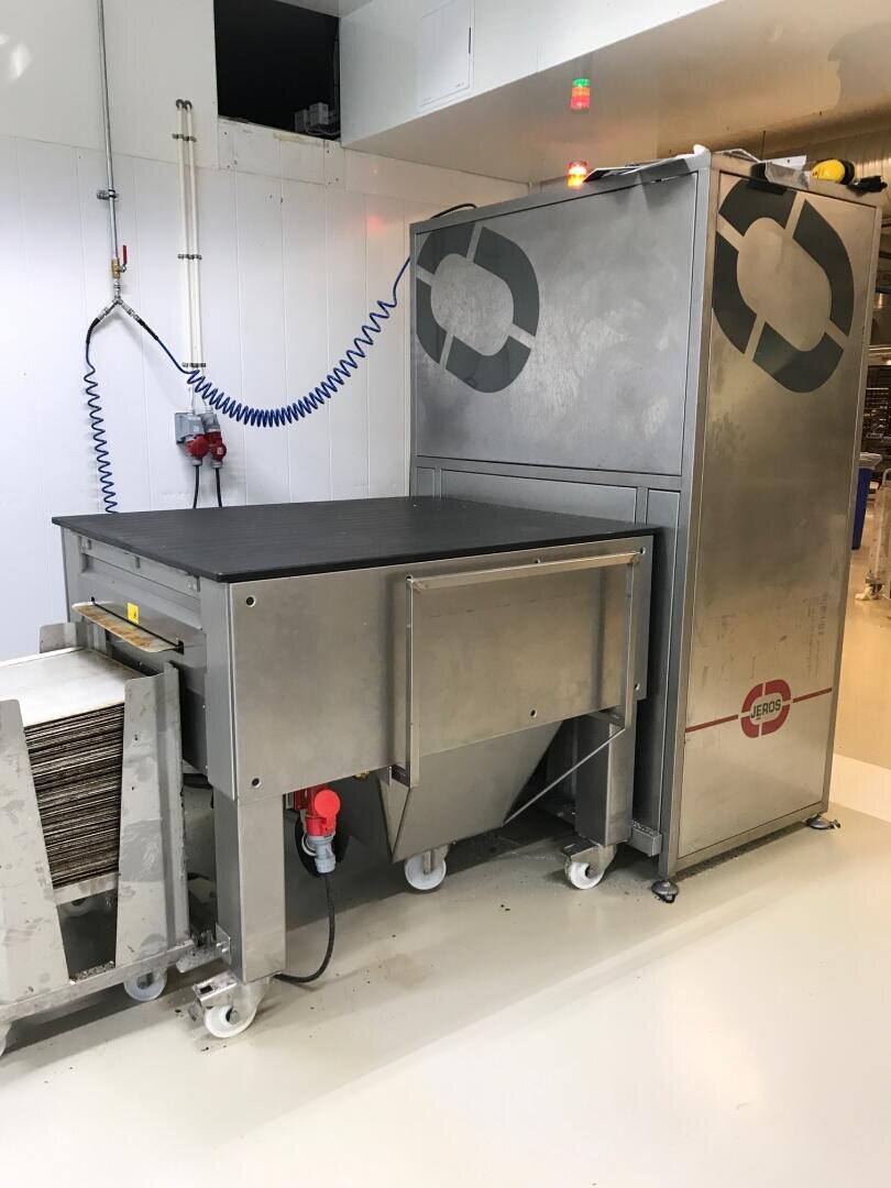 Eine große Industriemaschine aus rostfreiem Stahl mit Rädern und Bedienfeldern steht in einem sauberen, weißen Raum. Daneben sind ein blauer Schlauch und ein Stapel von Tabletts oder Platten zu sehen.