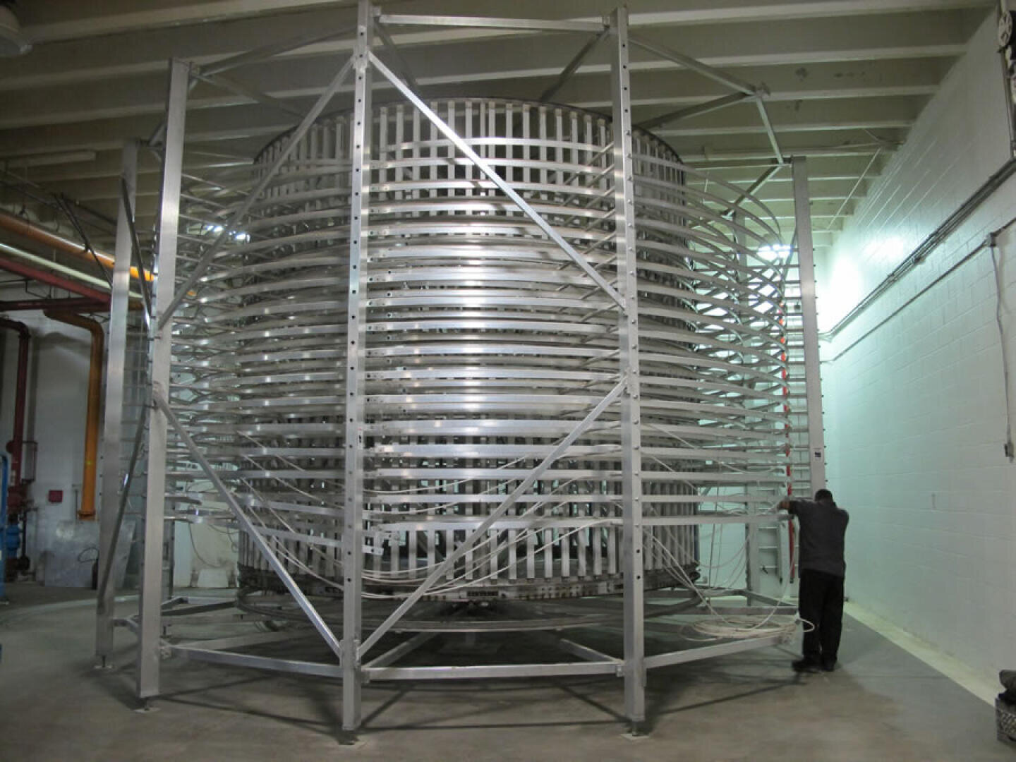 A large, cylindrical, metallic spiral chiller system stands in an industrial room, supported by a metal frame. A person is inspecting or working on the structure near its base. The room has white walls and overhead lighting.