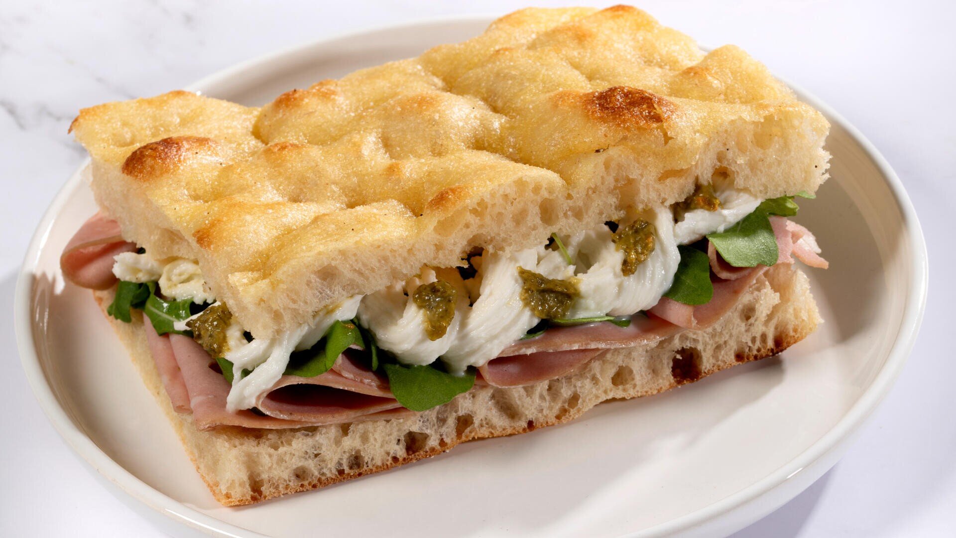 Das Sandwich auf einem weißen Teller besteht aus fluffigem Focaccia-Brot, das mit Schinken, frischem Mozzarella, Rucola und grünem Pesto belegt ist.