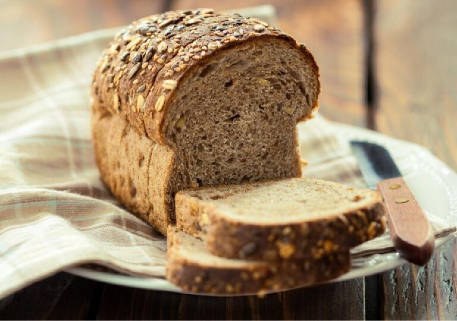 A loaf of multigrain bread with seeds on top sits on a plate, partially sliced. Two slices are cut and lie in front of the loaf, with a knife resting nearby on a plaid cloth.